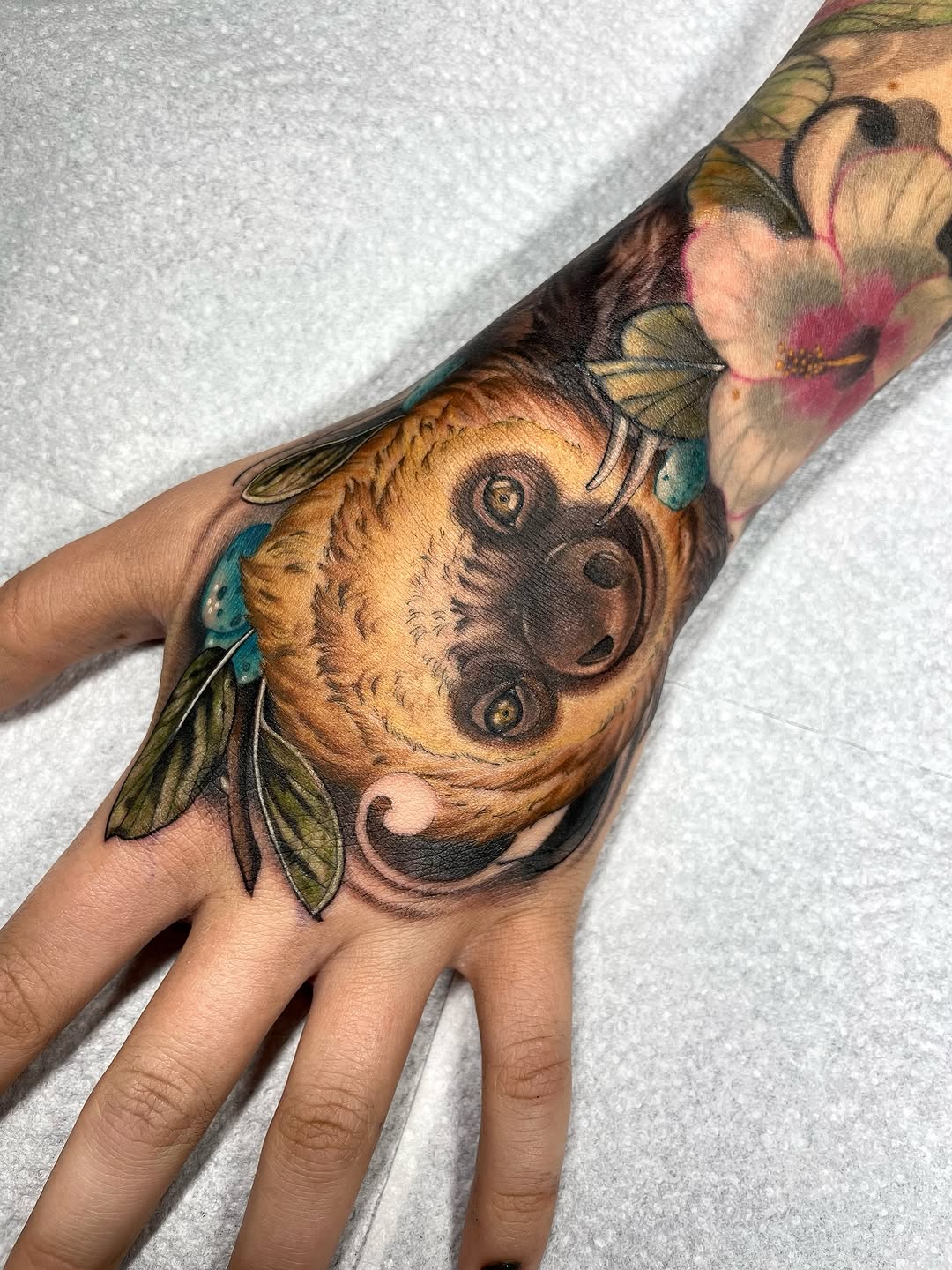 Colorful hand tattoo featuring an owl with yellow and brown tones, pink flowers, and blue butterfly wings extending up the forearm.