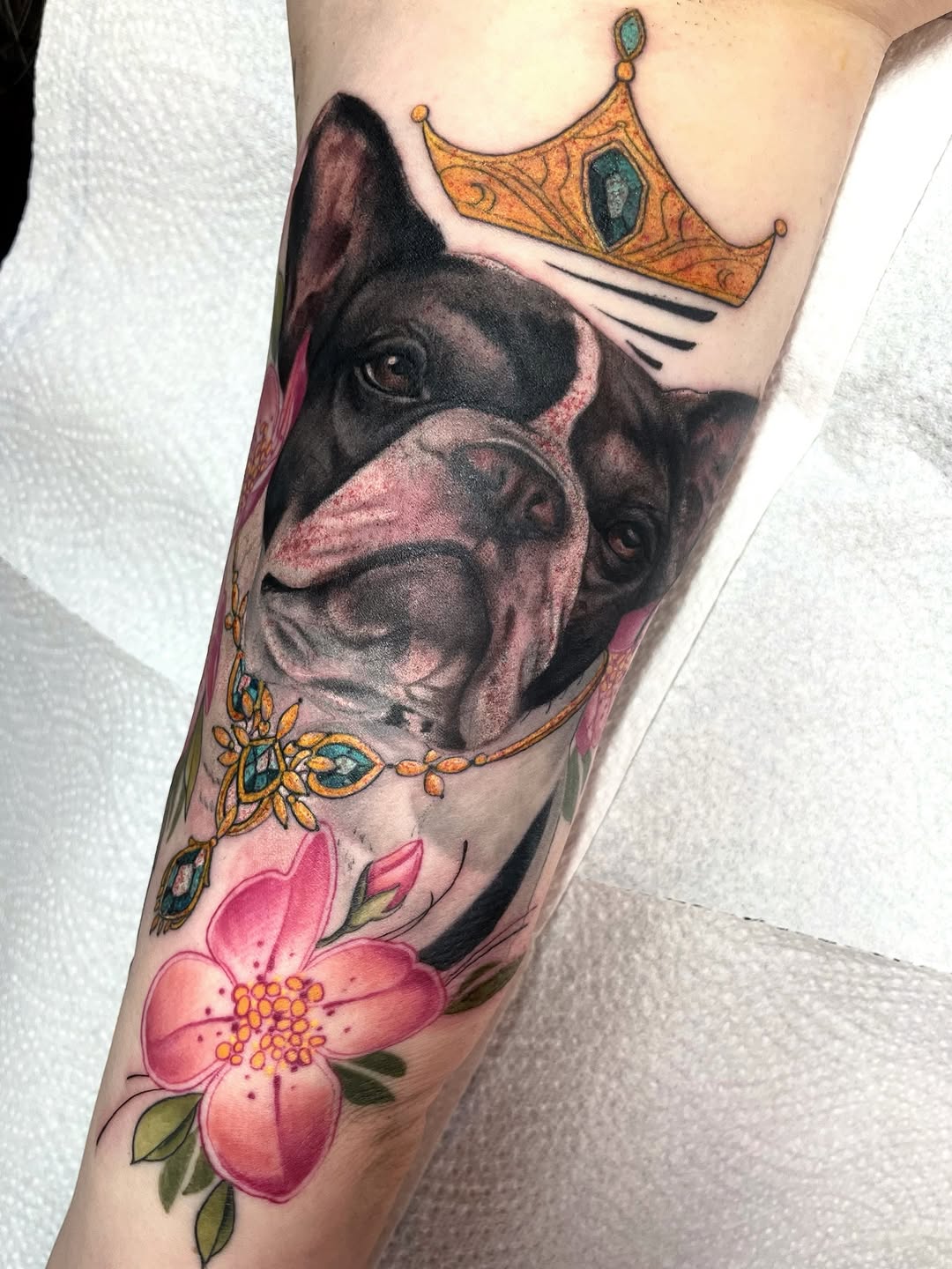 Colored tattoo on an arm featuring a dog wearing a crown, ornamental jewelry, pink flowers, and decorative gold elements.