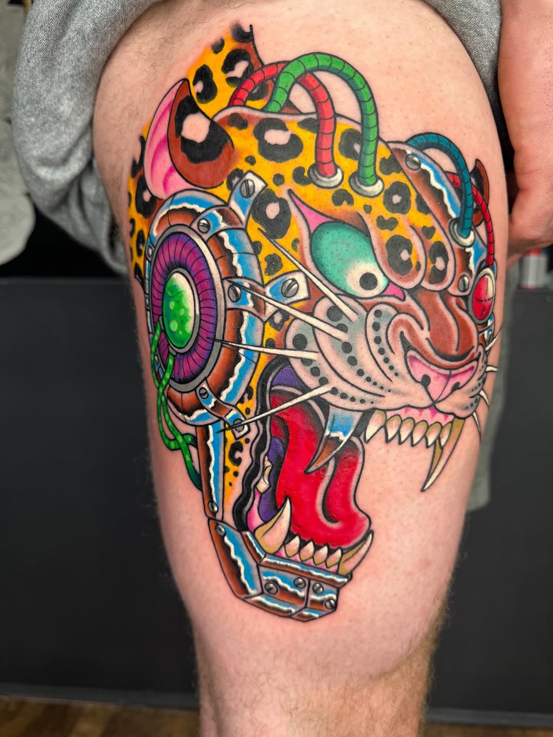 Colorful skull tattoo on thigh featuring leopard spots, mechanical elements, green snake, and bright yellow, pink, red, and blue details.