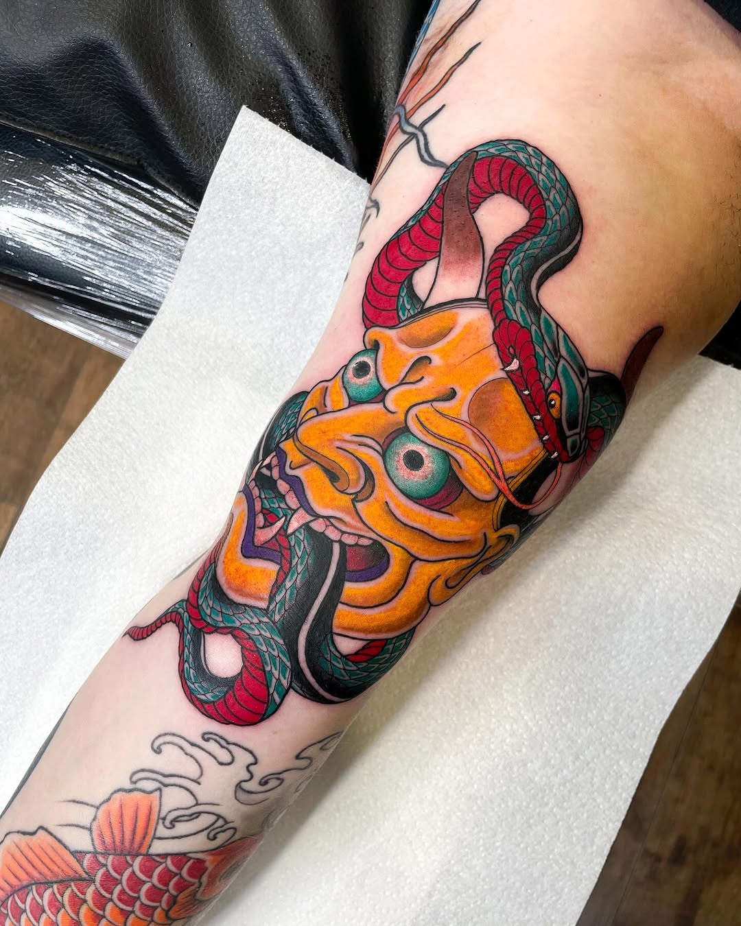 Colorful tattoo on the forearm featuring an orange demon face with green eyes and red serpents, with additional fish imagery visible below.