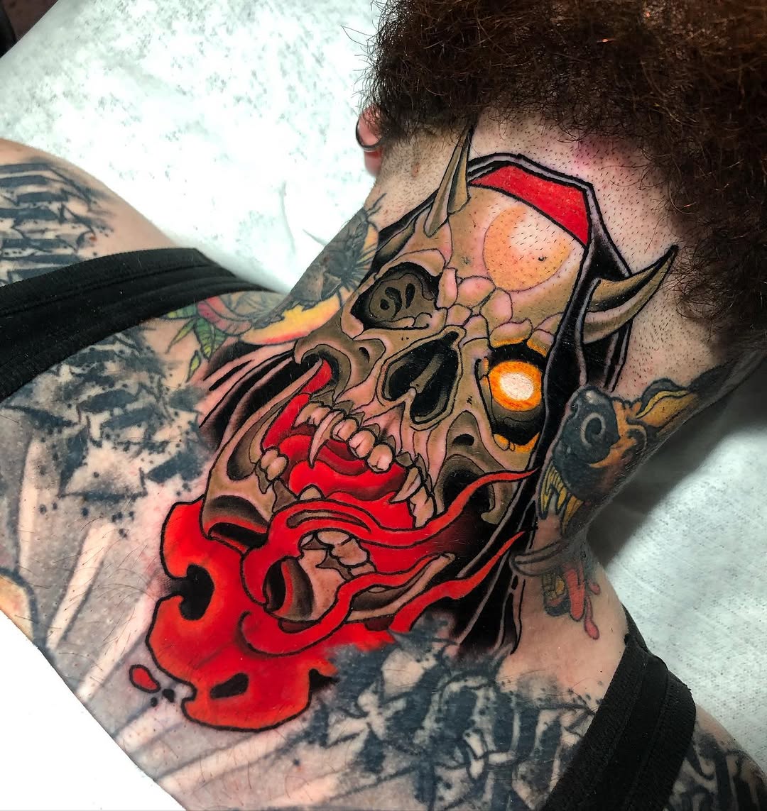 Colorful demon skull head tattoo on neck and shoulder with red flames, orange eyes, and black horns against skin with surrounding dark ink work.