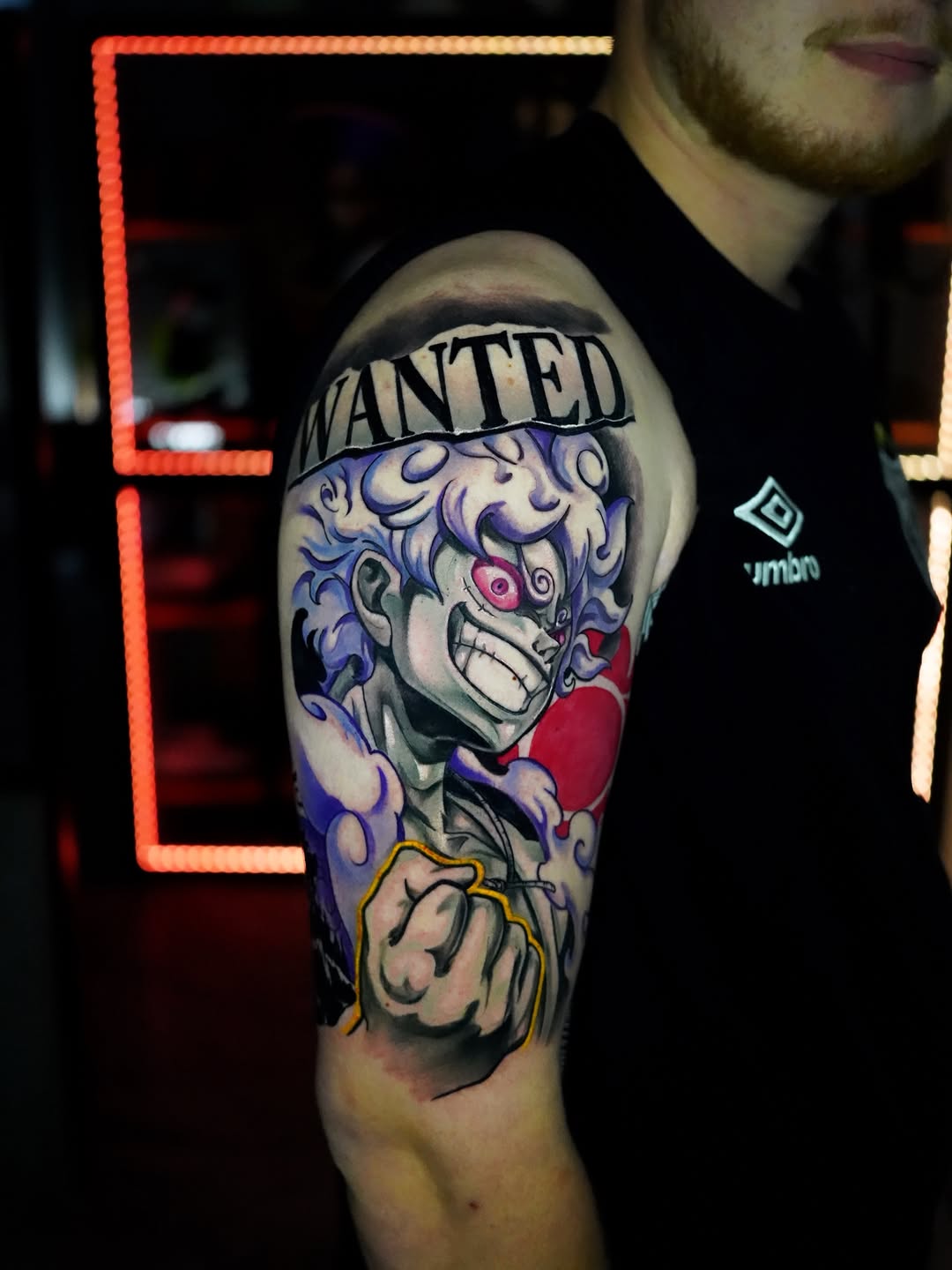 Colorful shoulder tattoo featuring a skull with purple hair, "WANTED" banner, red heart, and grinning expression in blue, red, and yellow tones.