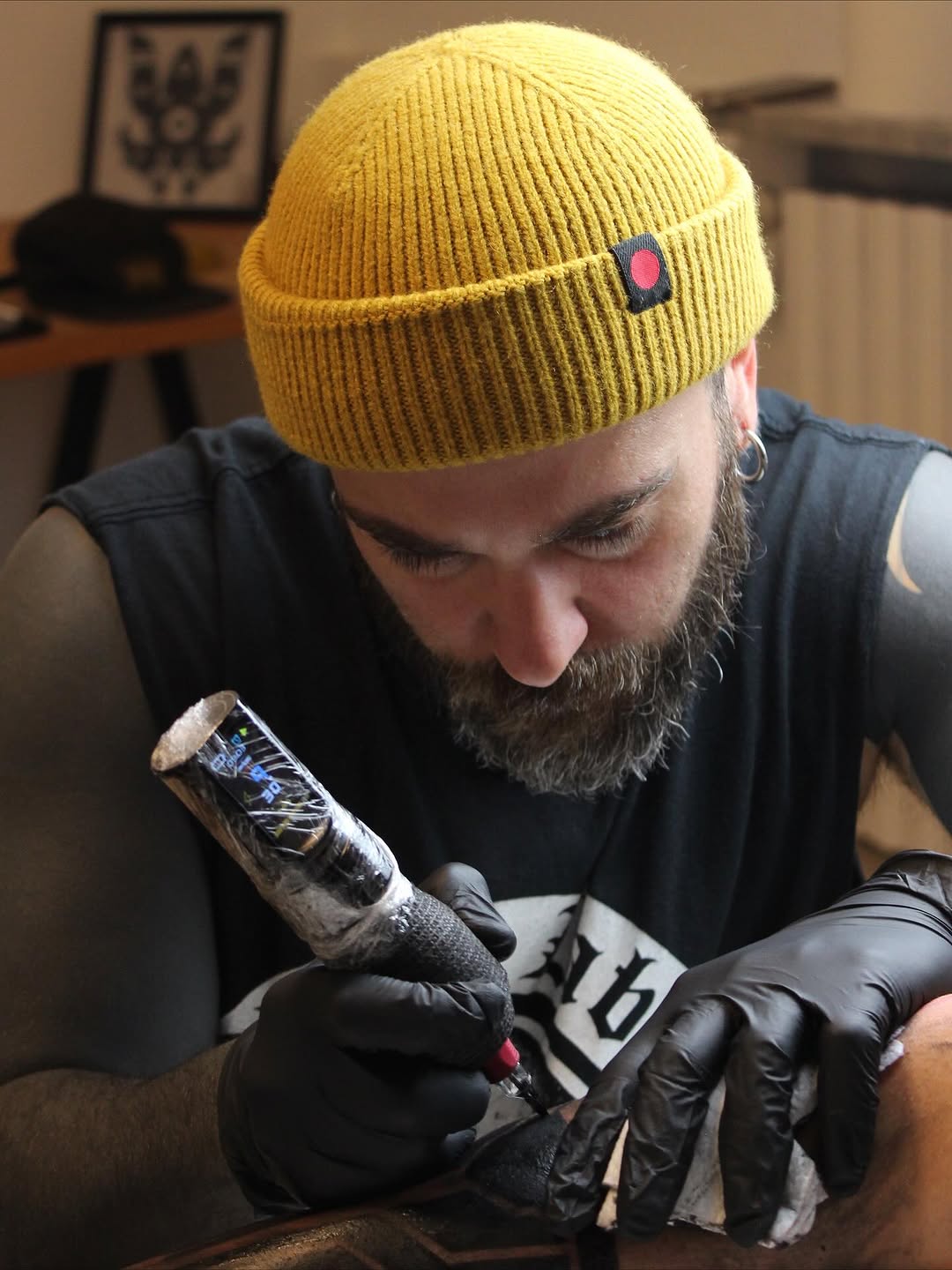 Bearded tattoo artist wearing a yellow beanie and black gloves examines a fresh tattoo on a client's arm featuring pink and blue colors.