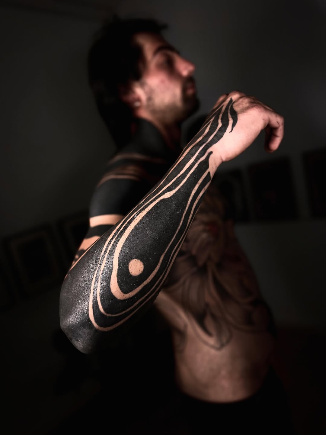 Man displaying a large black and skin-tone tattoo on his forearm featuring curved lines and circular elements in a geometric design.