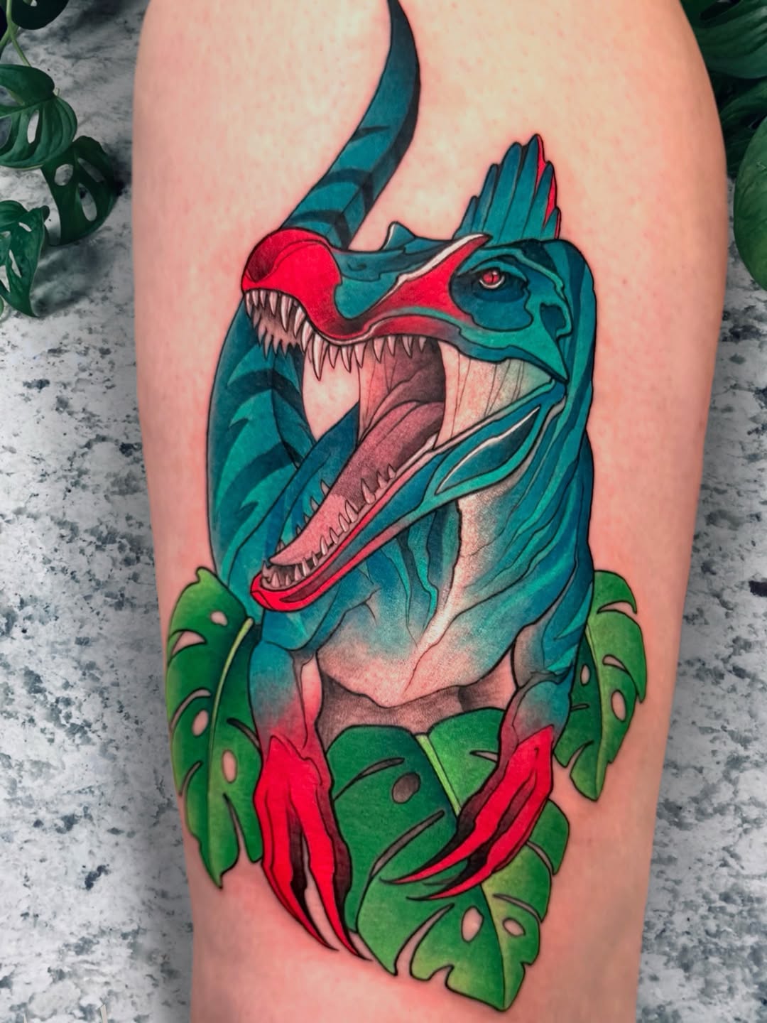 Colorful dinosaur head tattoo with open jaws in teal and red, surrounded by green tropical leaves on the upper leg.