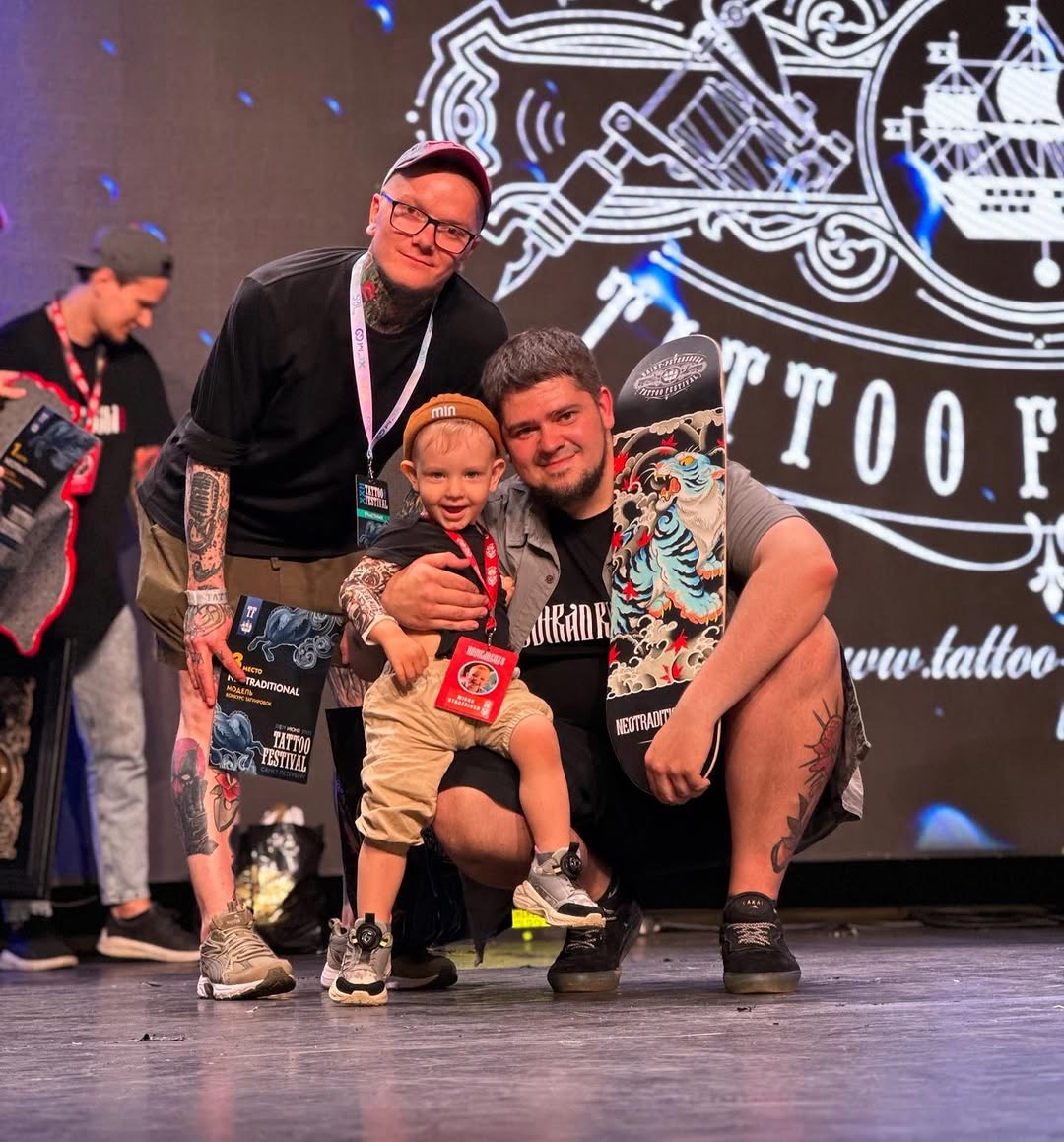 Three men posing on stage at a tattoo festival, with visible arm and chest tattoos featuring colorful designs including tigers and ornamental elements.