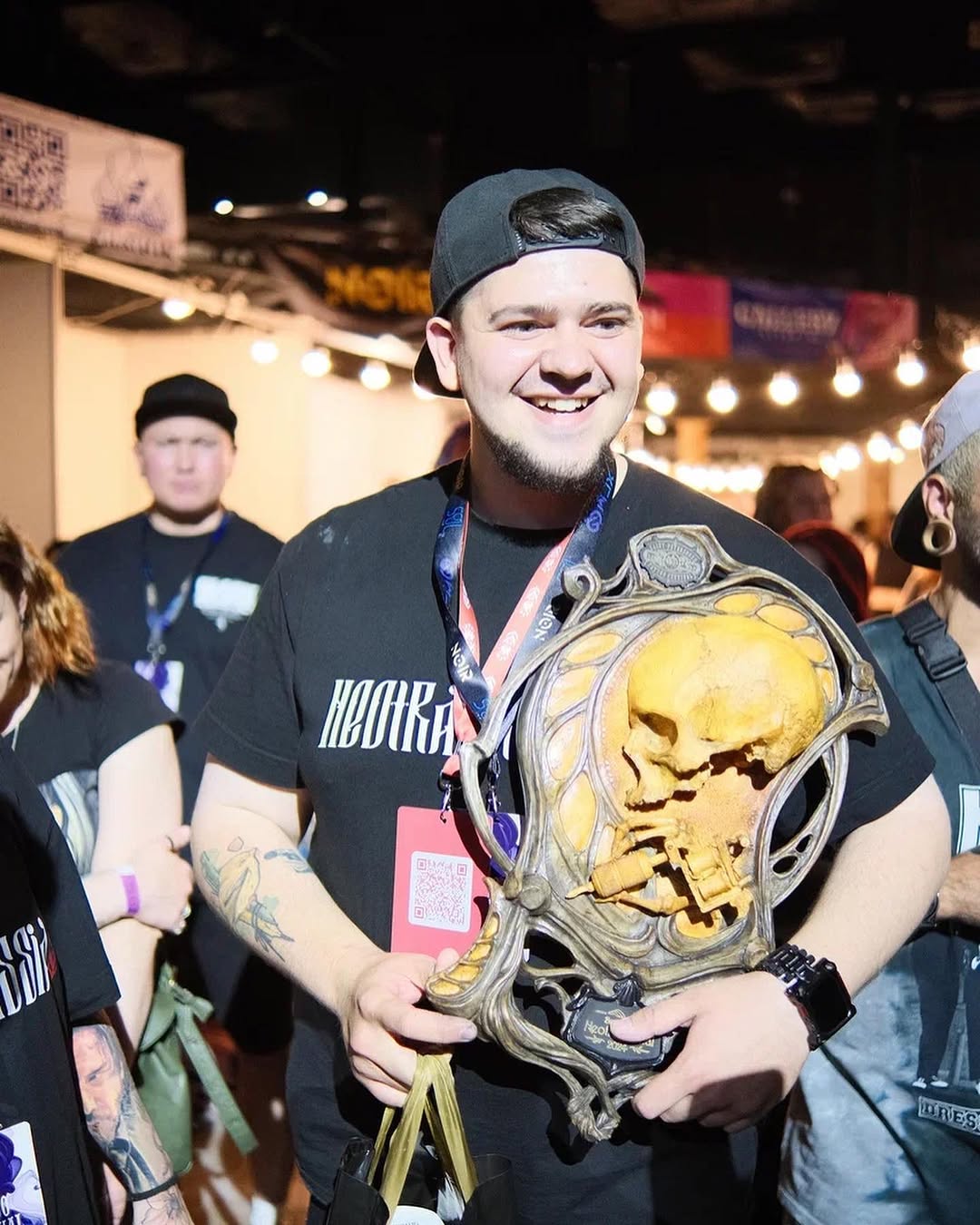 Smiling man wearing a blue cap and black shirt displays a large golden skull tattoo on his upper arm at an indoor event.