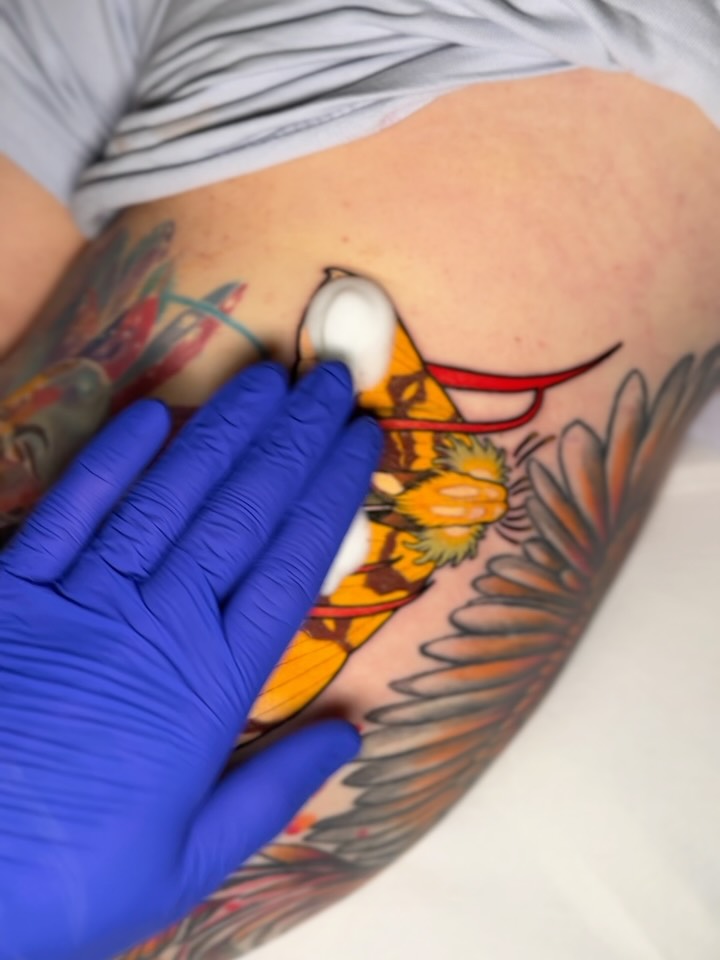 Colorful bird tattoo on the upper arm featuring yellow, red, orange, and black ink, with a gloved hand holding the skin during application.