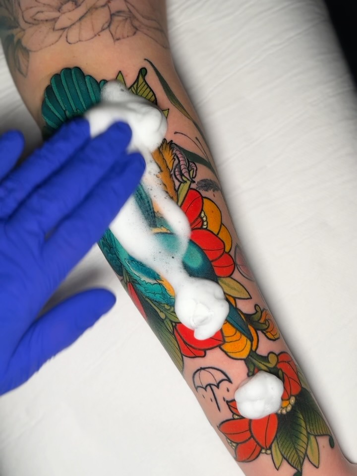 Colorful floral tattoo on forearm featuring red, orange, and teal flowers with green leaves, being cleaned with a white cloth by a gloved hand.