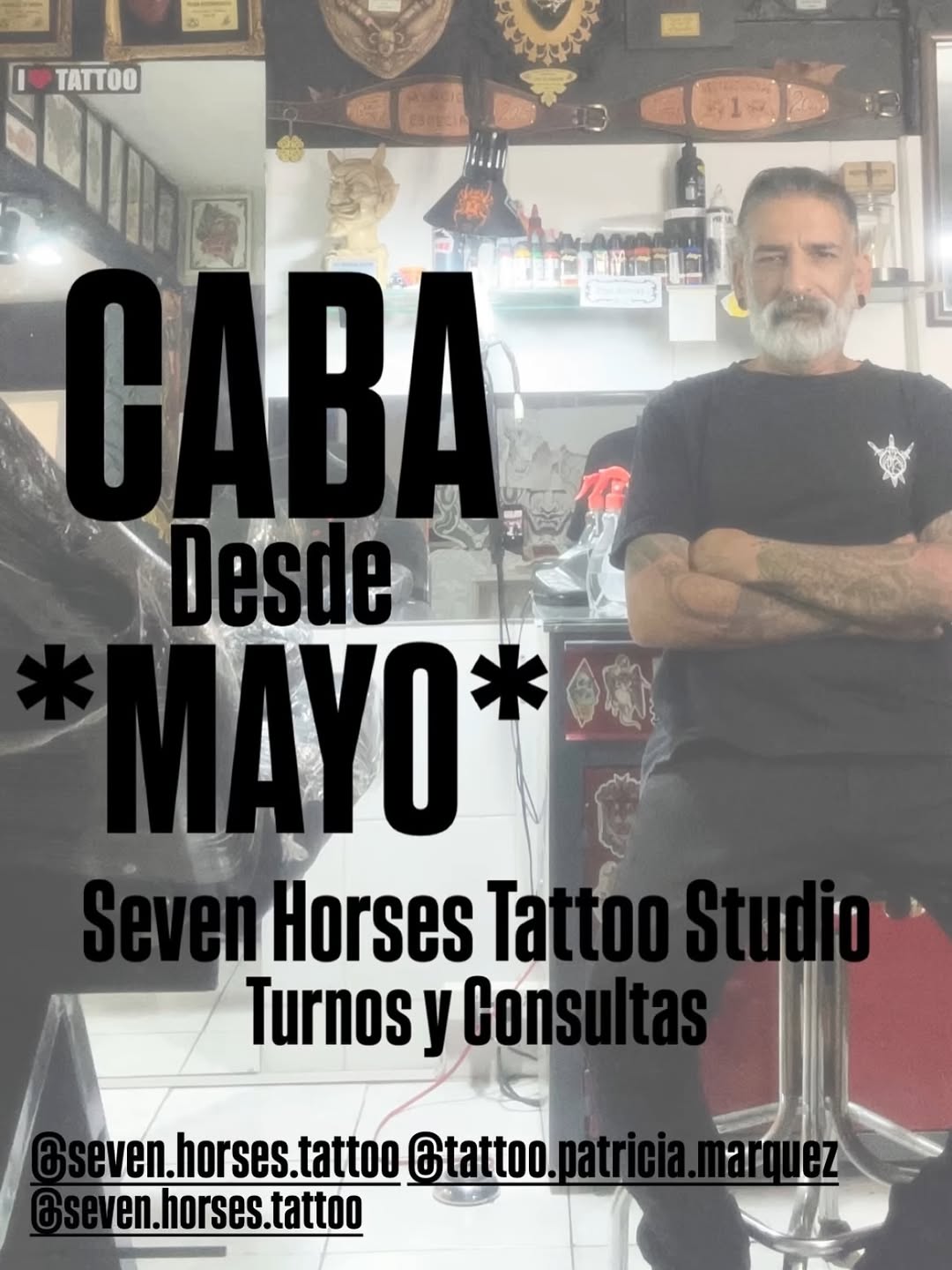 Tattoo artist with beard standing in studio wearing black shirt with ship wheel logo, surrounded by ink bottles and studio decorations.