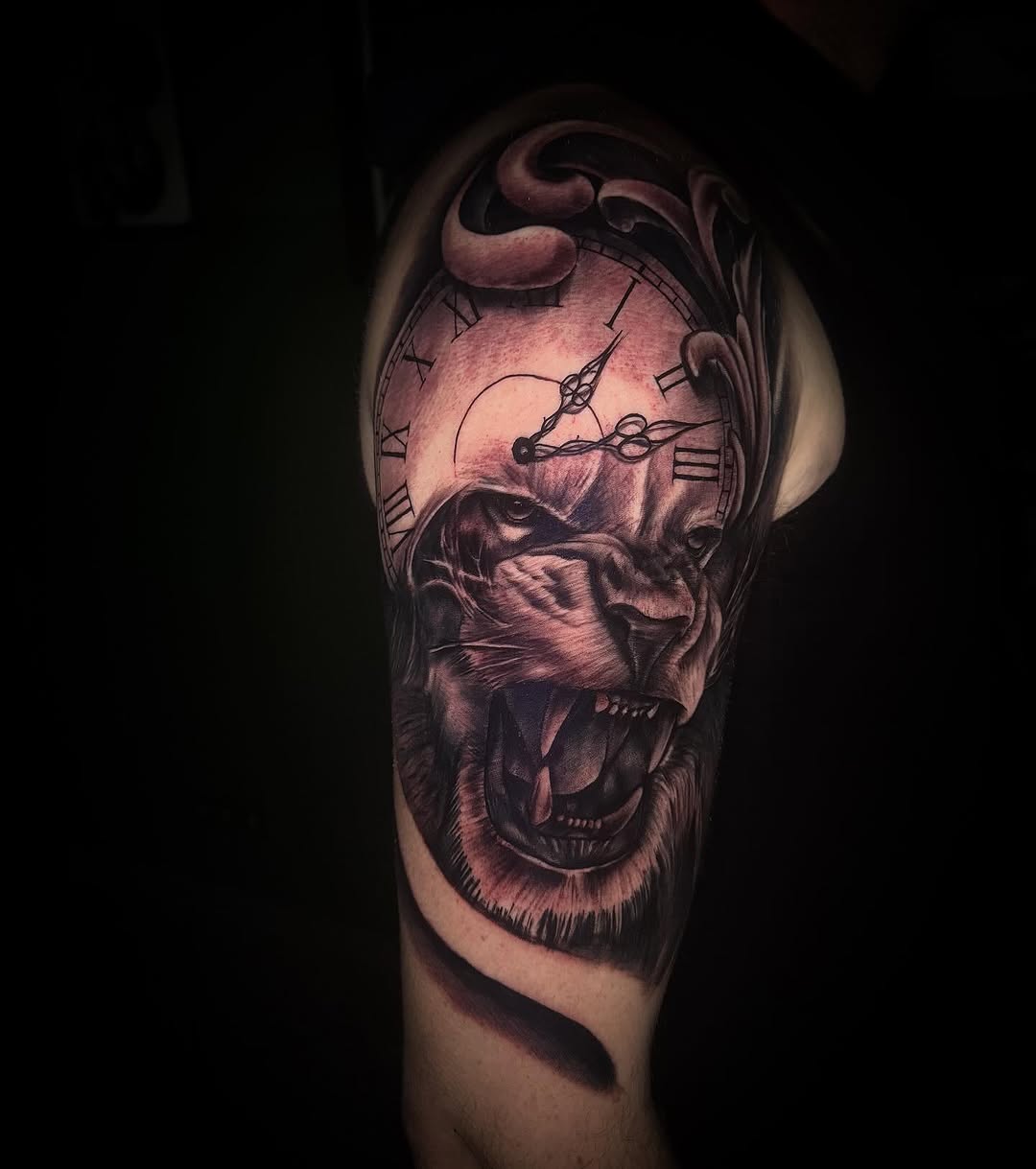 Detailed upper arm tattoo featuring a bearded male face with an open mouth expression and a clock face integrated into the forehead, rendered in red and black tones.