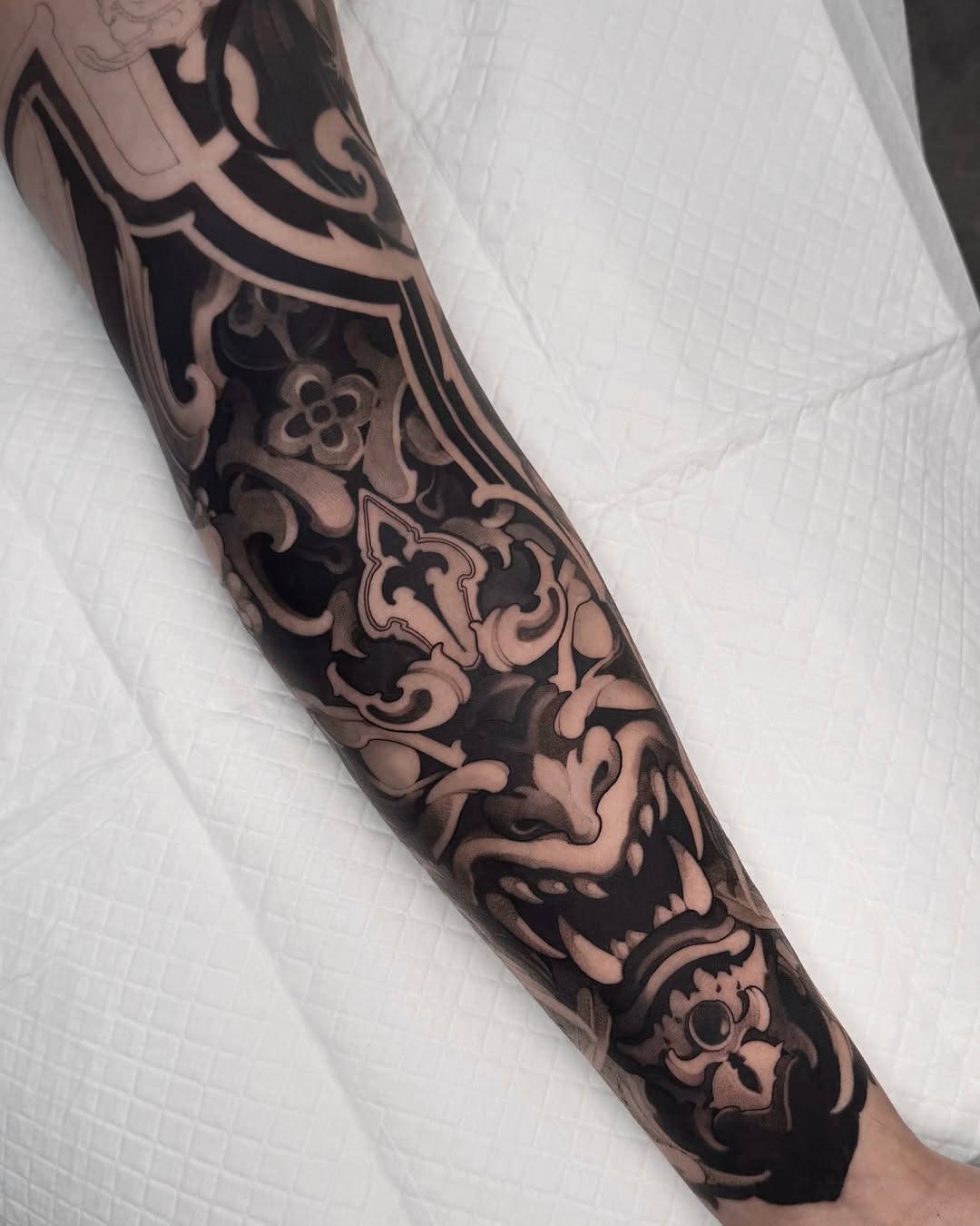 Ornate decorative sleeve tattoo featuring scrolling flourishes, floral motifs, and a central face design in black and grey tones on the forearm.