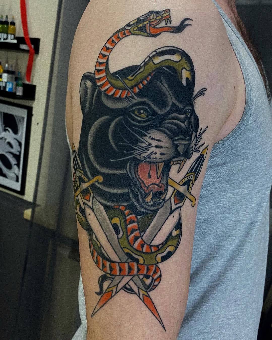 Panther head with open mouth surrounded by two intertwined snakes with orange and black stripes, daggers, and spikes on upper arm.