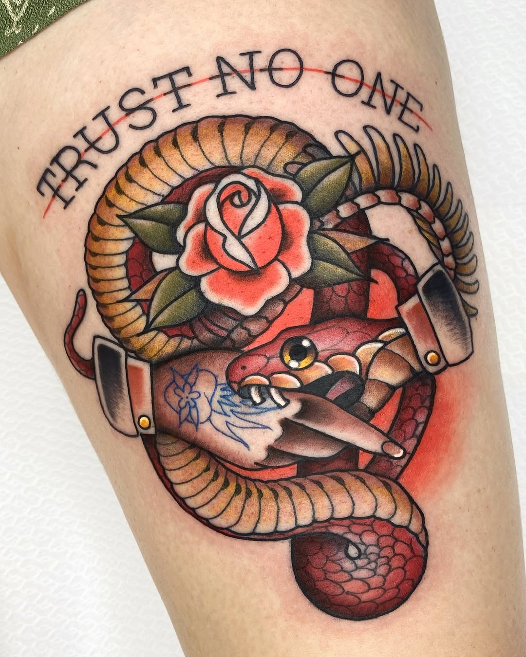 Colorful thigh tattoo featuring a coiled snake with horns, red rose, hands holding a knife, and text reading "Trust no one" in an arc.