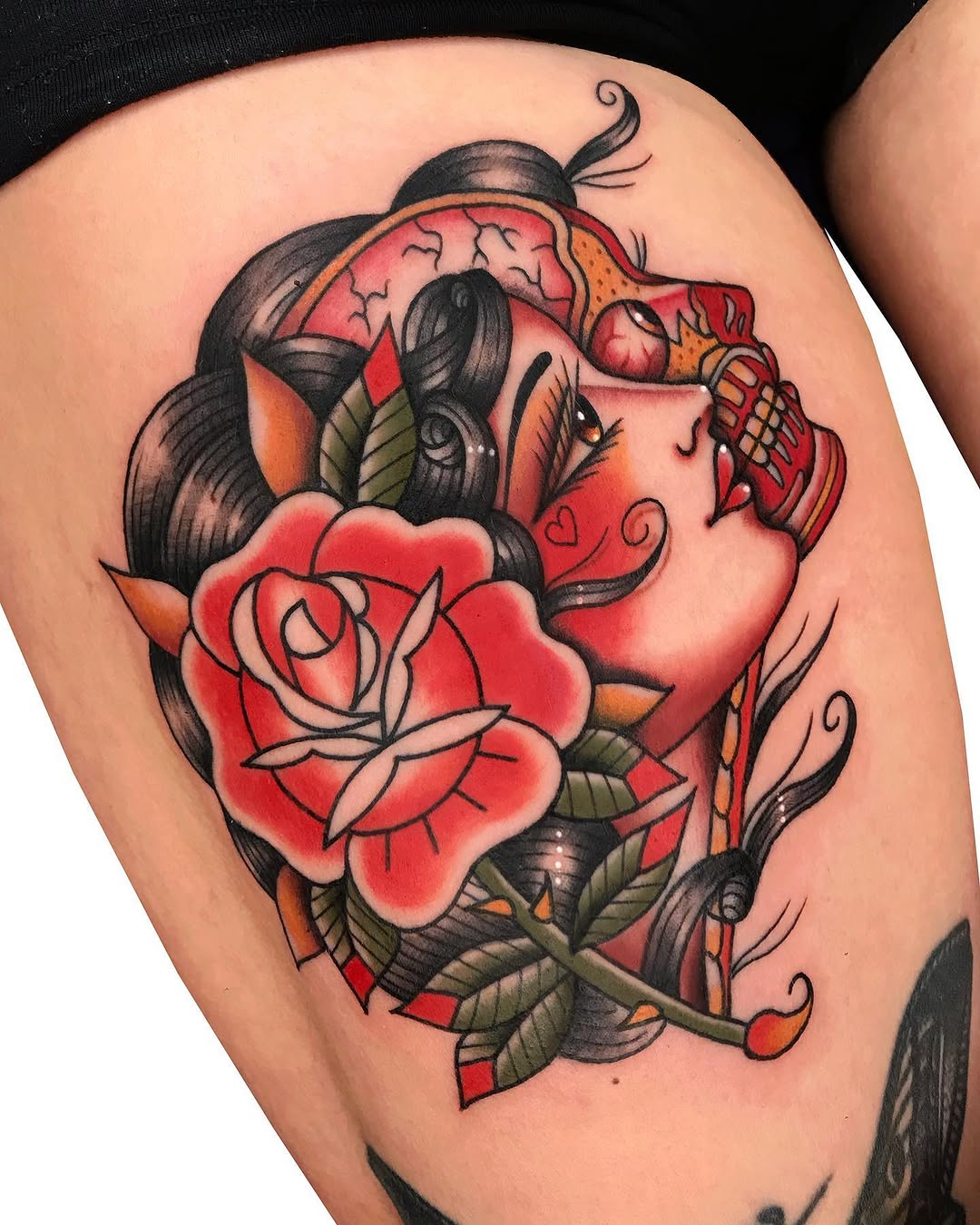 Colorful tattoo on upper thigh featuring a woman's face with dark hair, red rose, ladybug, and decorative swirls in red, orange, and black ink.
