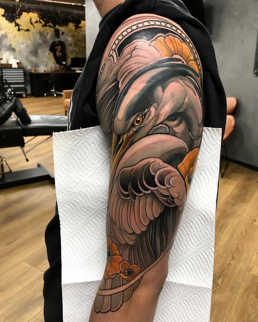 Detailed eagle head tattoo on upper arm featuring gray and orange coloring, ornamental headband, and feathered wings extending down the sleeve.
