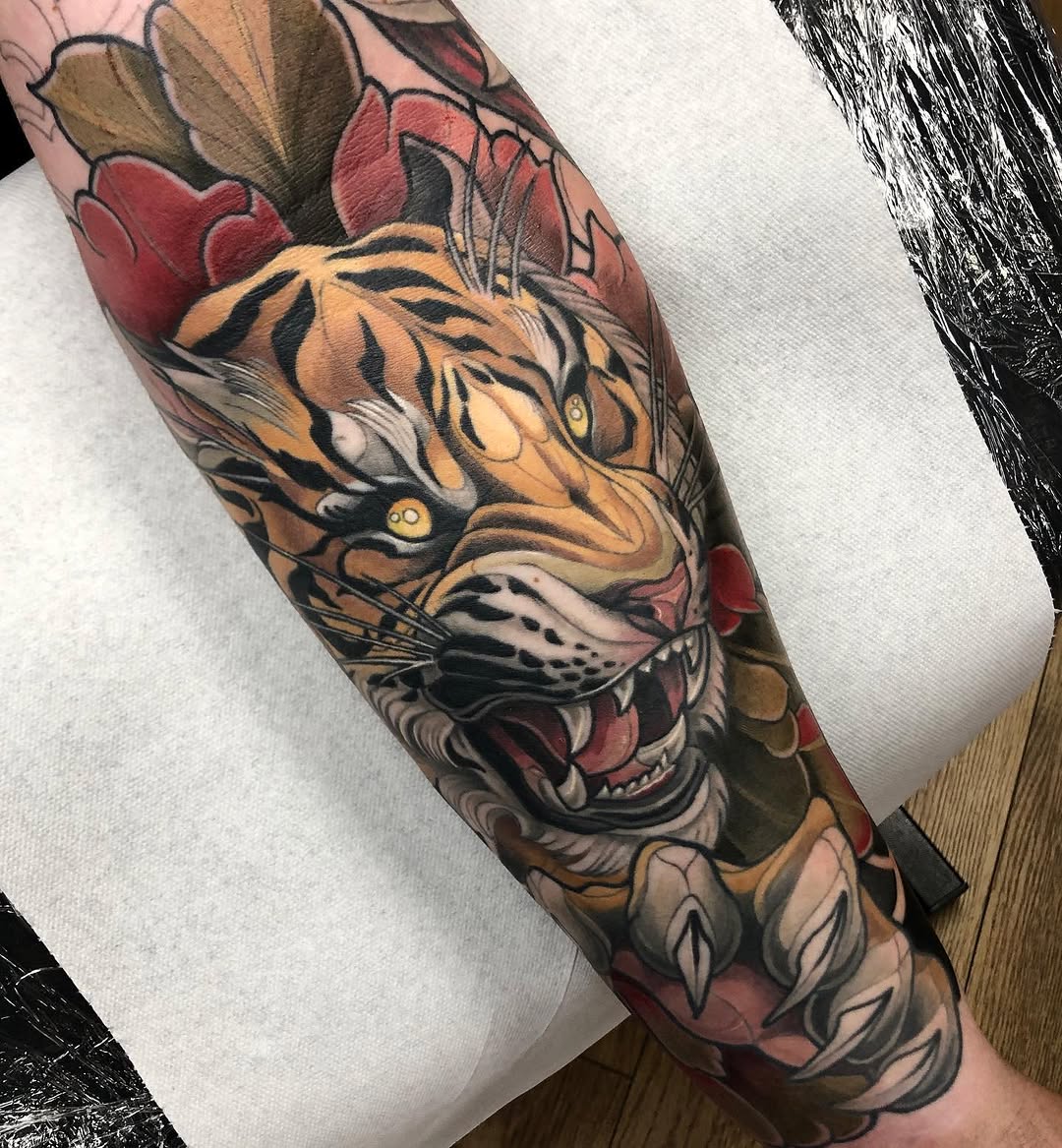 Tiger head with bared teeth and yellow eyes surrounded by red and burgundy flowers on a forearm sleeve.
