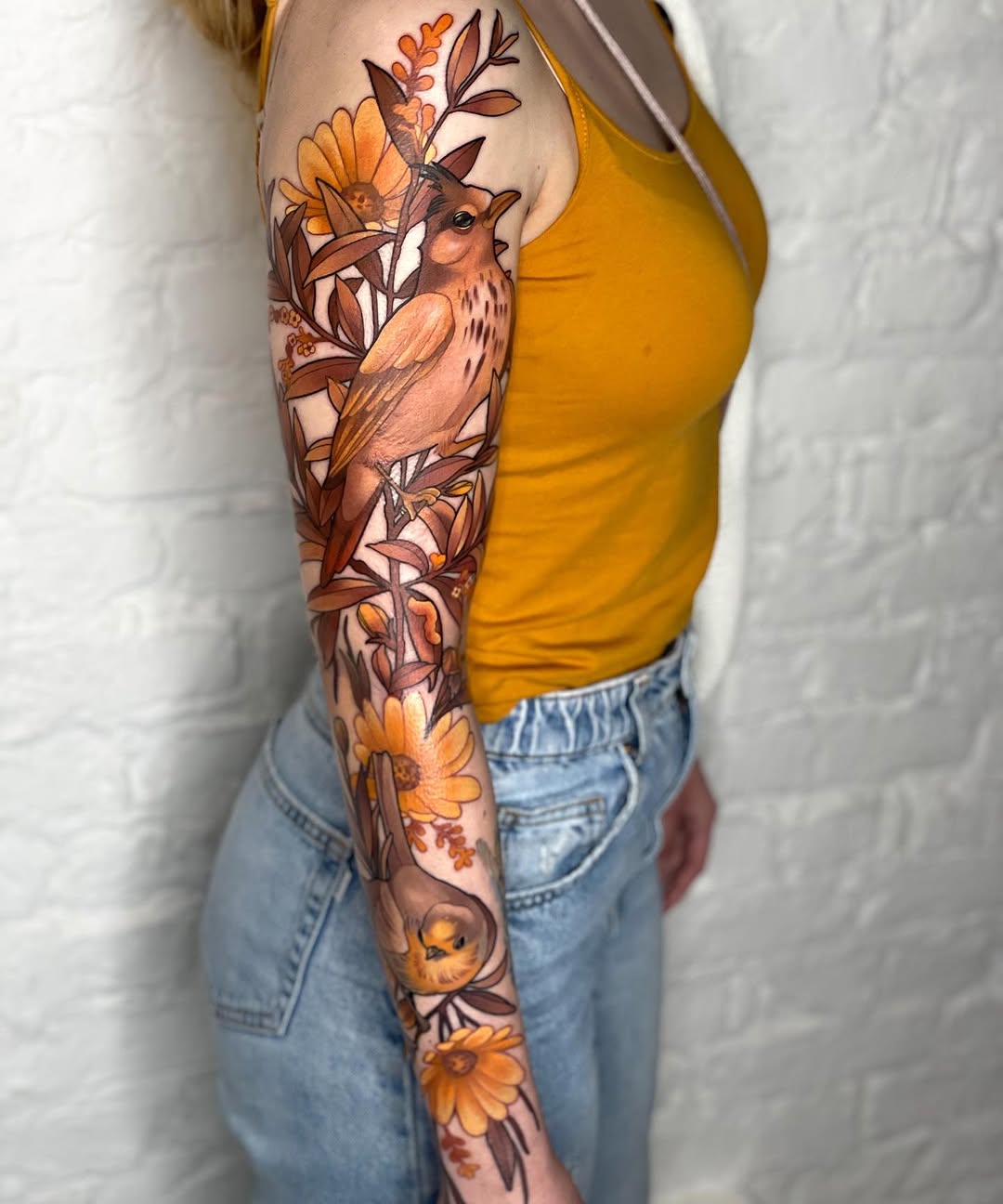 Full-sleeve arm tattoo featuring an owl surrounded by sunflowers, daisies, and botanical leaves in warm orange, brown, and yellow tones.