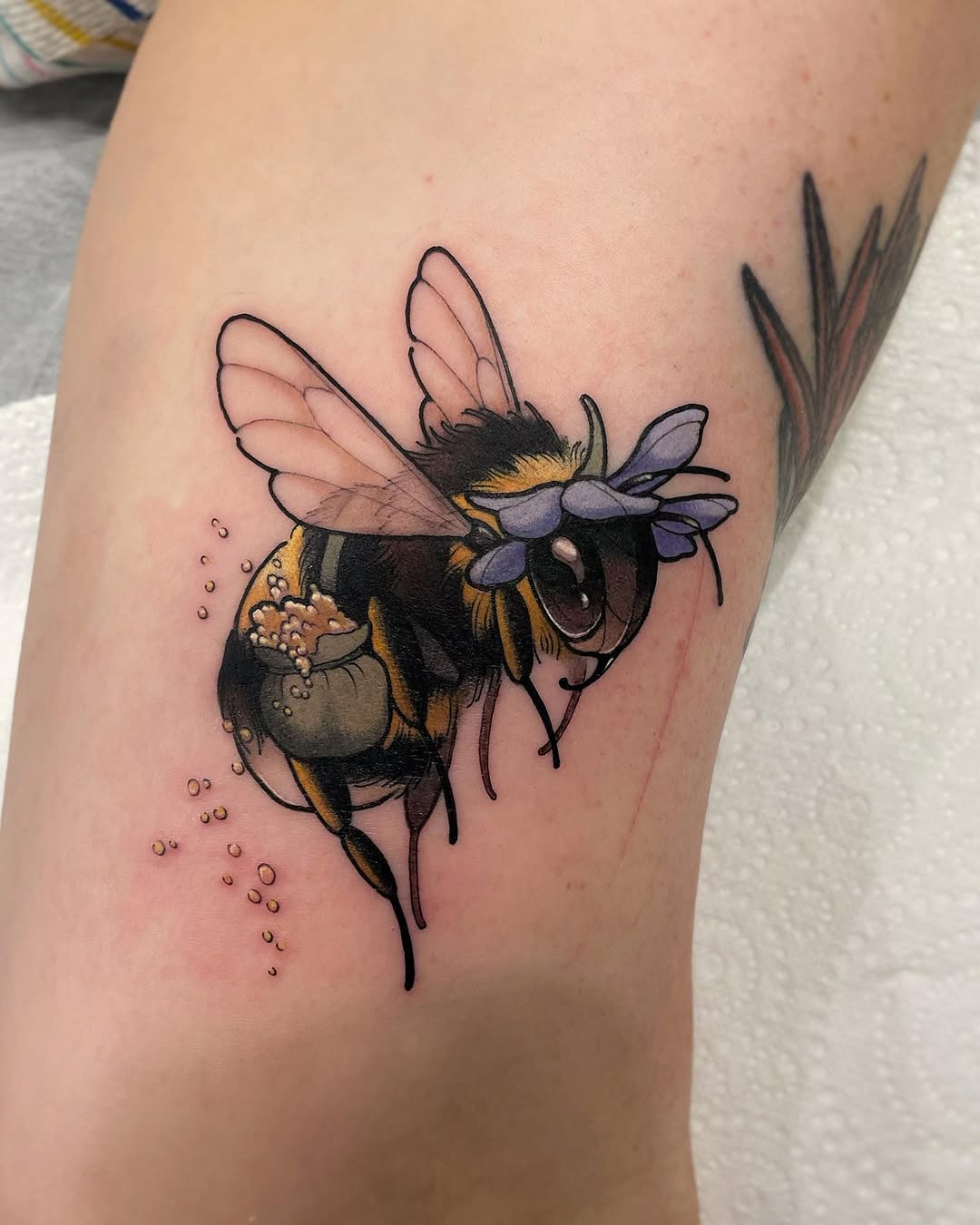 Colorful bee tattoo on thigh carrying a pollen sac, with purple flowers and scattered pollen droplets trailing below.