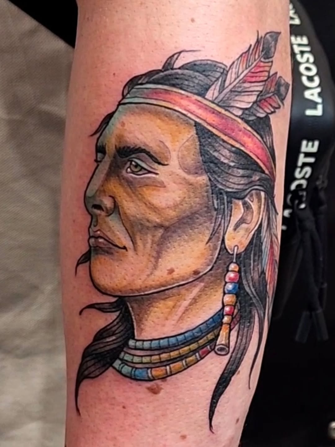 Colorful portrait tattoo of a Native American man in profile wearing a feathered headdress and beaded necklace on the forearm.