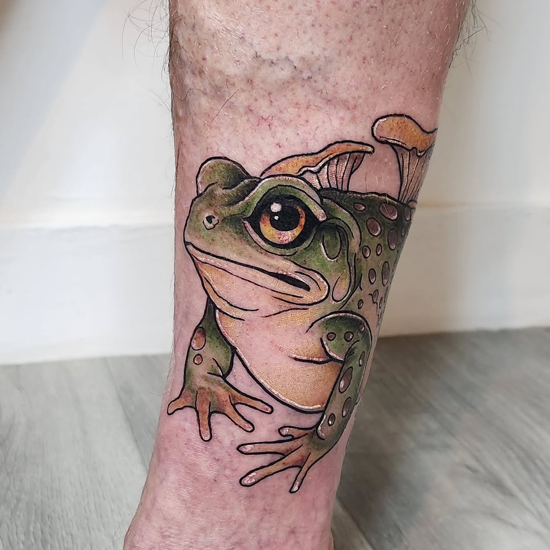 Colorful frog tattoo on the calf featuring green, tan, and orange tones with detailed eyes and spotted patterns.