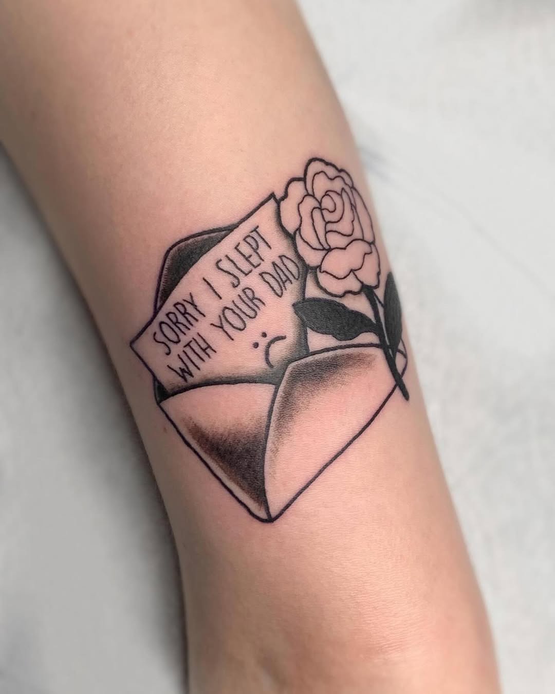 Envelope tattoo with rose and handwritten text reading "Sorry I slept with your dad" on inner arm with black ink and shading.