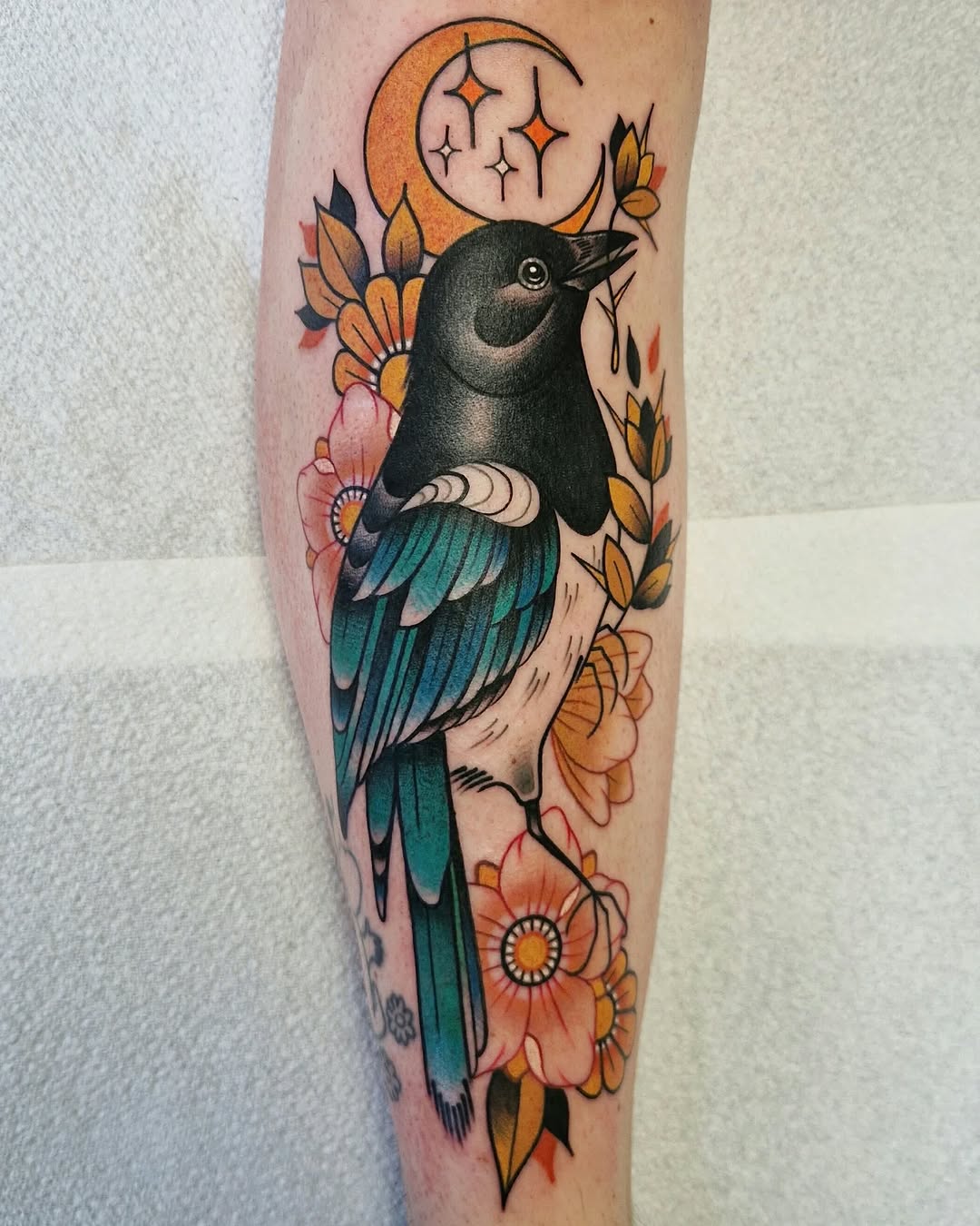 Black crow with teal wings perched among orange and pink flowers with a crescent moon above, tattooed on the forearm.
