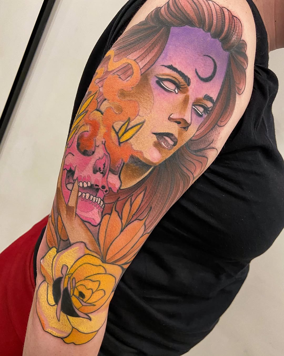 Colorful upper arm tattoo featuring a woman's face in purple and pink with a skull overlay, yellow roses, and orange floral elements.