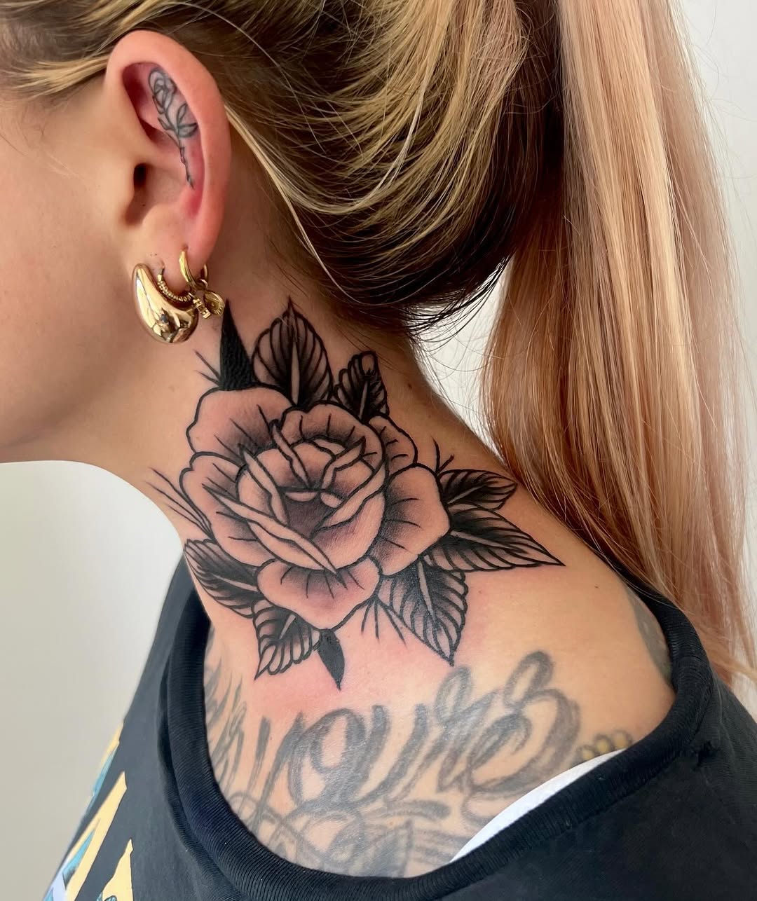 Rose tattoo with dark leaves on the neck and shoulder of a person with blonde hair wearing gold hoop earrings and a black top.