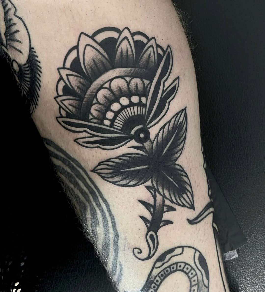 Black ink flower tattoo on upper arm with layered petals, decorative center details, leafy stem, and ornamental elements below.