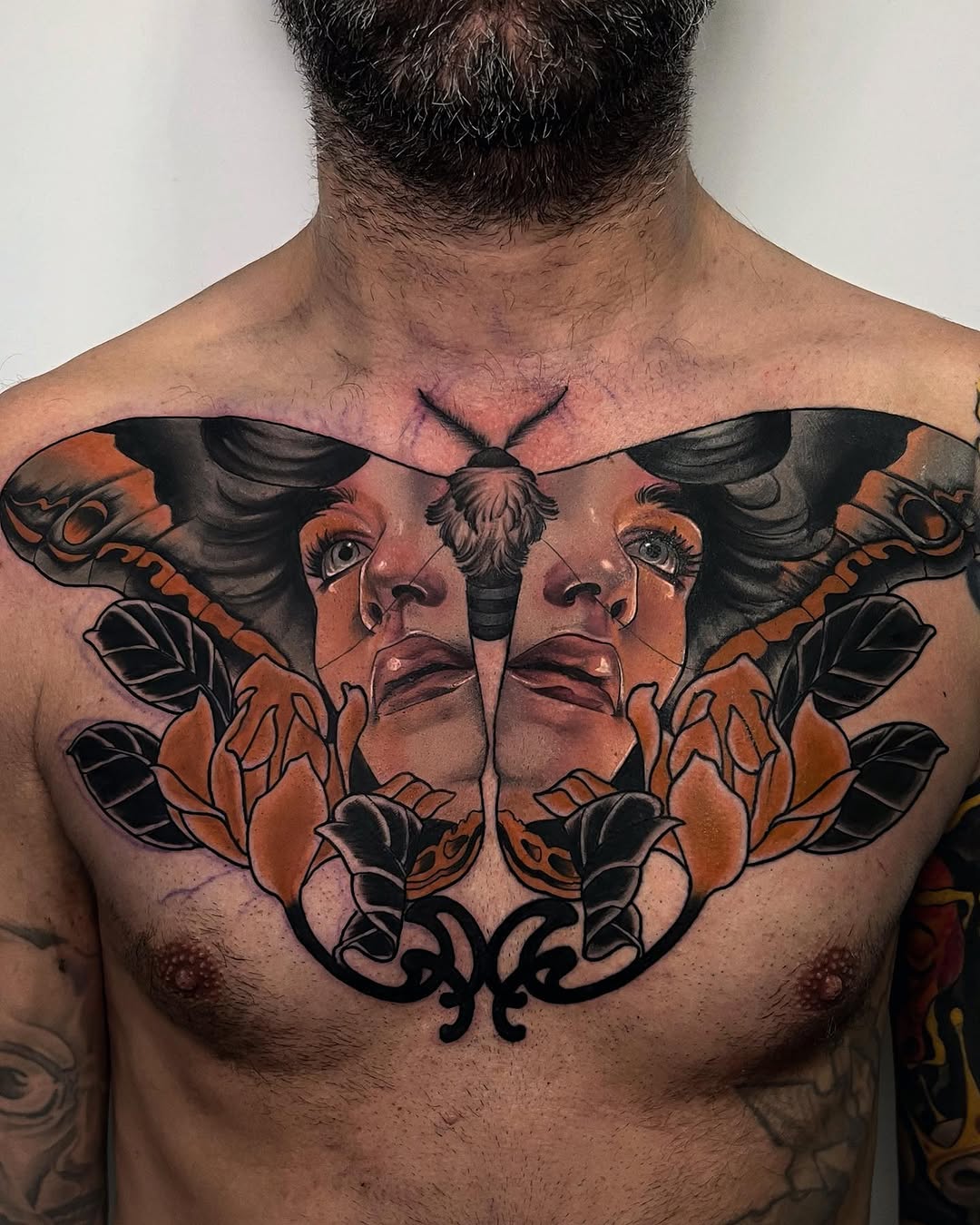 Symmetrical chest tattoo featuring mirrored female faces with moth wings in orange and black ink, with ornamental flourishes below.