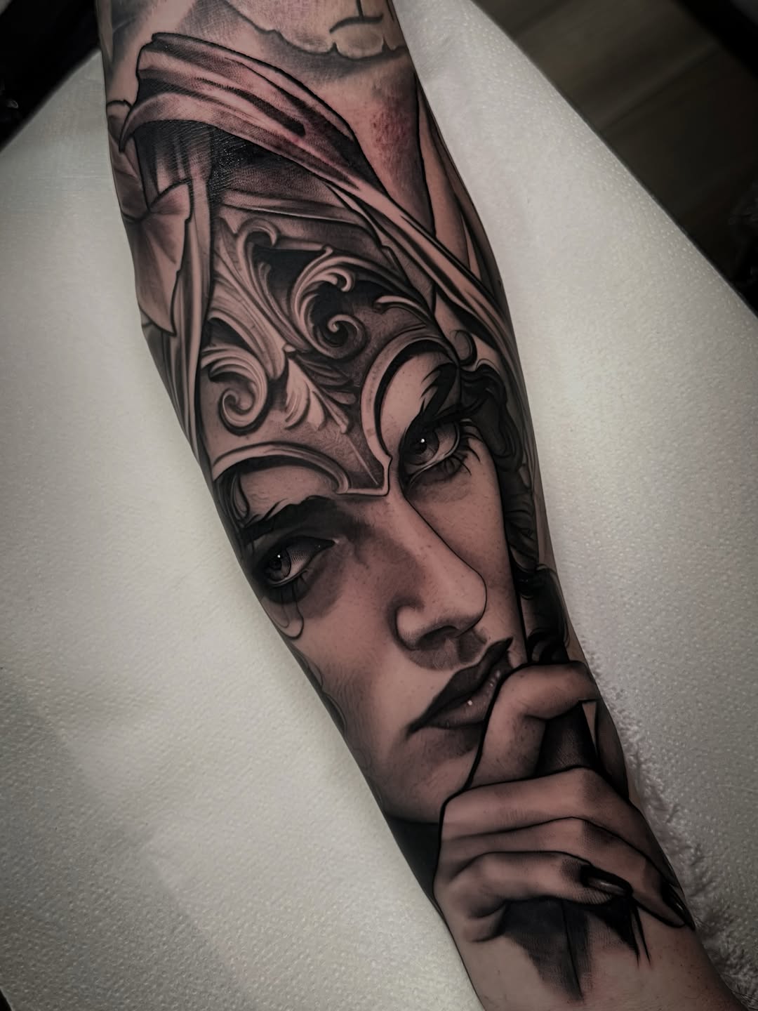 Portrait tattoo on forearm featuring a woman's face with ornate mask design, shaded in black and grey tones with decorative scrollwork.