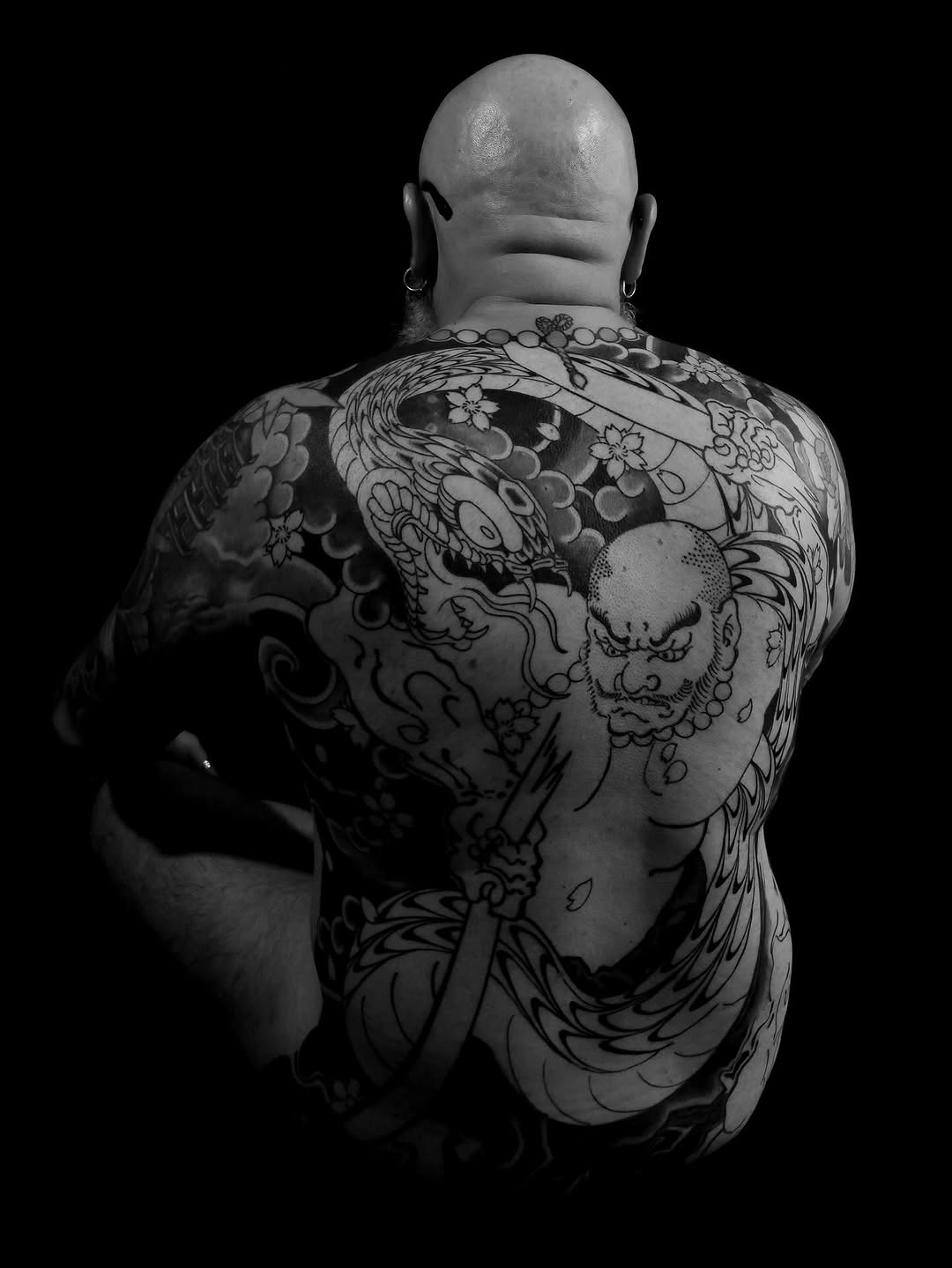 Rear view of a shirtless person displaying extensive back and shoulder tattoos featuring dragons, demon faces, and floral motifs in black ink on pale skin.