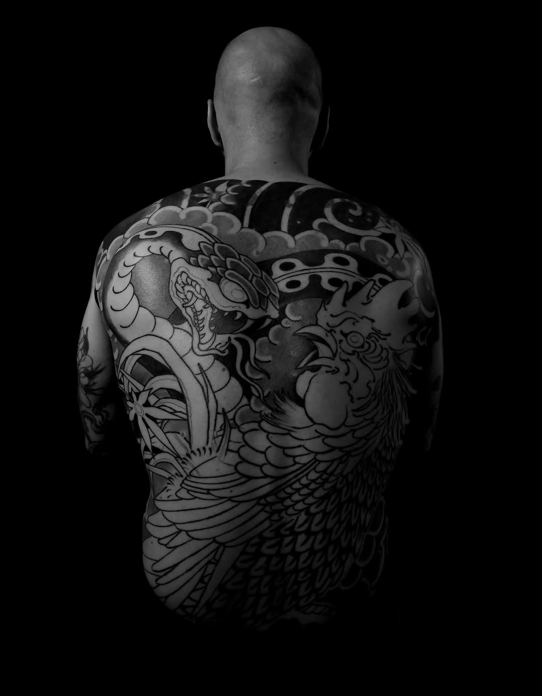 Rear view of a shirtless man with full back tattoo featuring dragons, clouds, and ornamental designs in black ink on pale skin against a dark background.