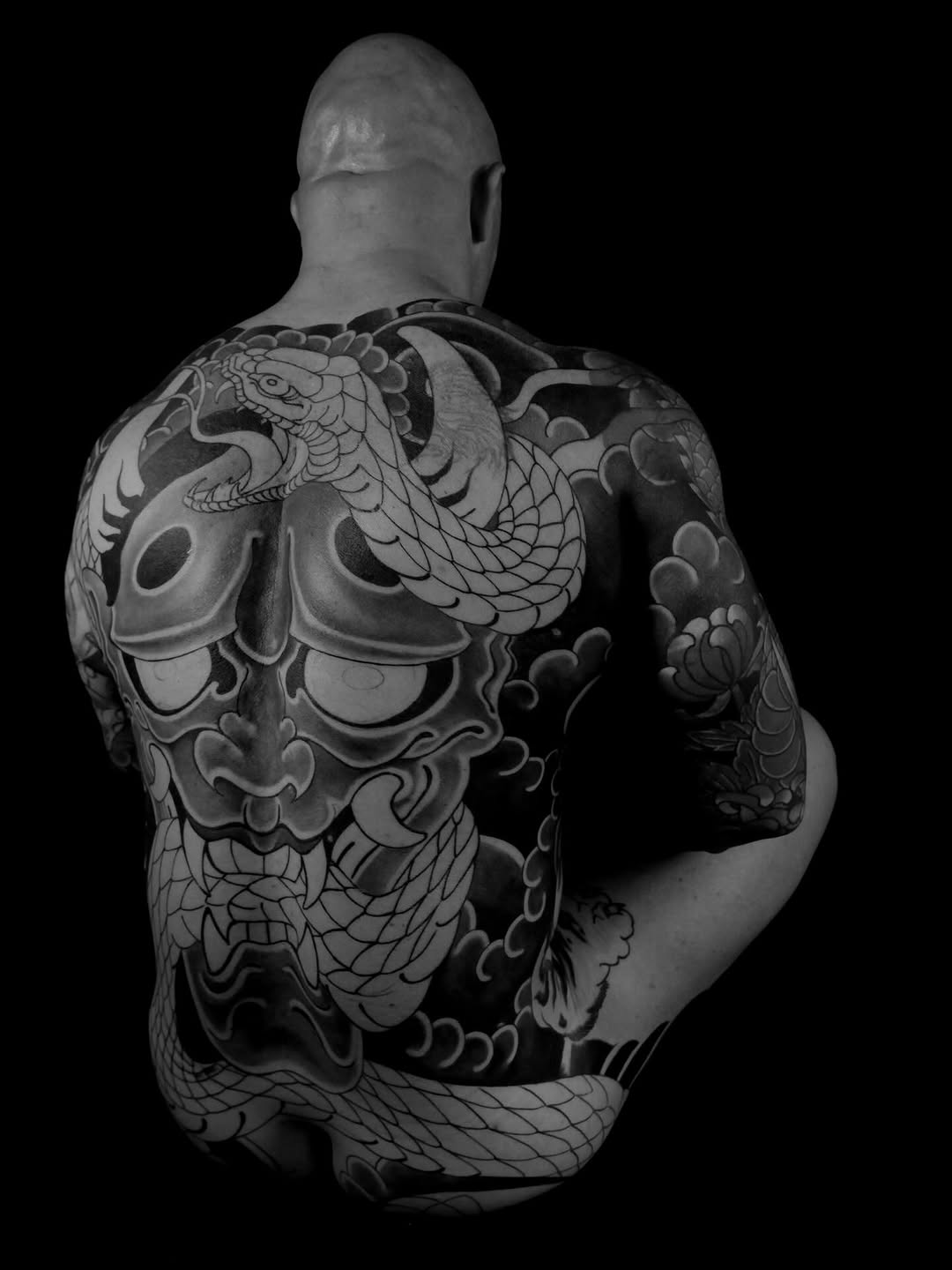 Back piece tattoo featuring dragons, ornamental masks, and flowing botanical elements in black ink on a shirtless man against a black background.