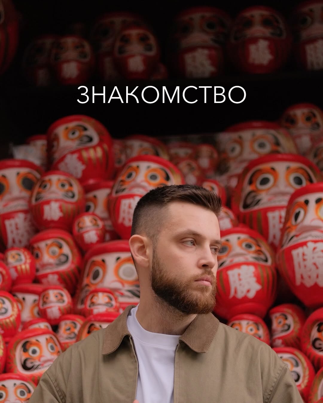 Man with a beard wearing a tan jacket and white shirt, positioned against a background of red and orange decorative dolls with painted faces.