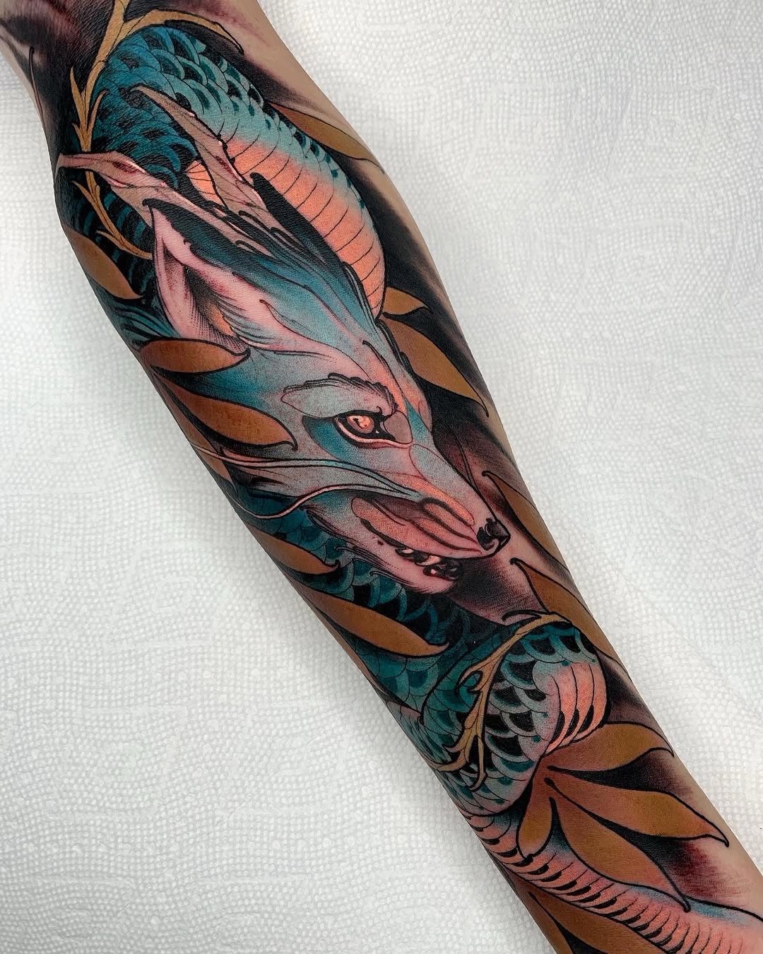 Colorful dragon and fox mask tattoo on forearm featuring teal, pink, gold, and bronze tones with scaled and feathered details.