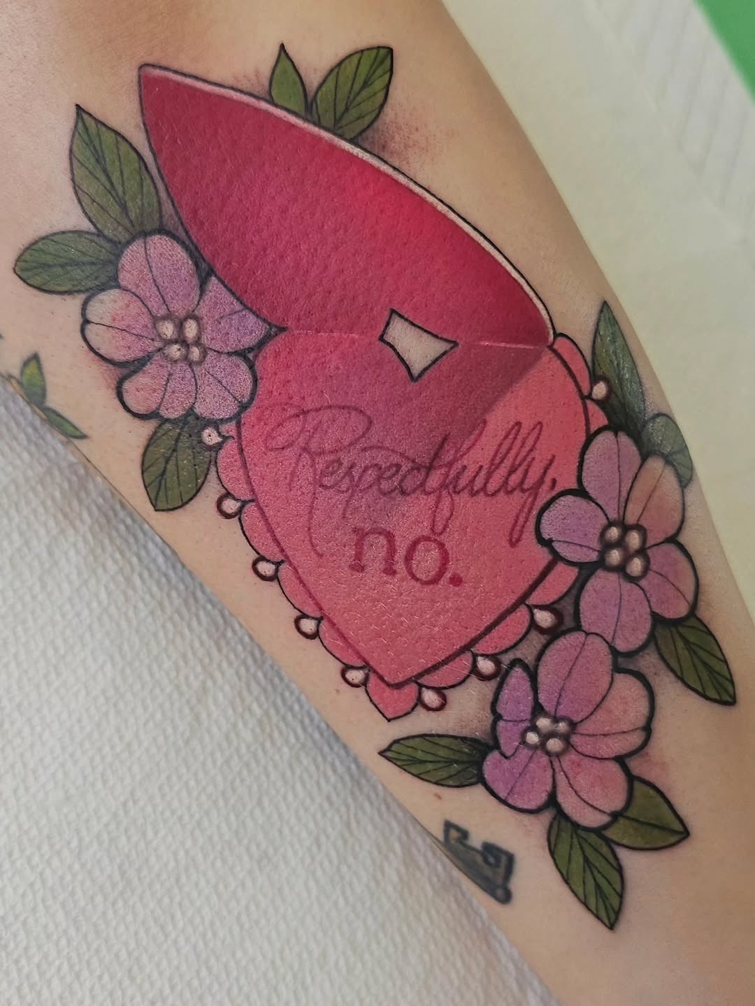 Red heart with script text surrounded by pink flowers and green leaves on an upper arm, with artist initials below.