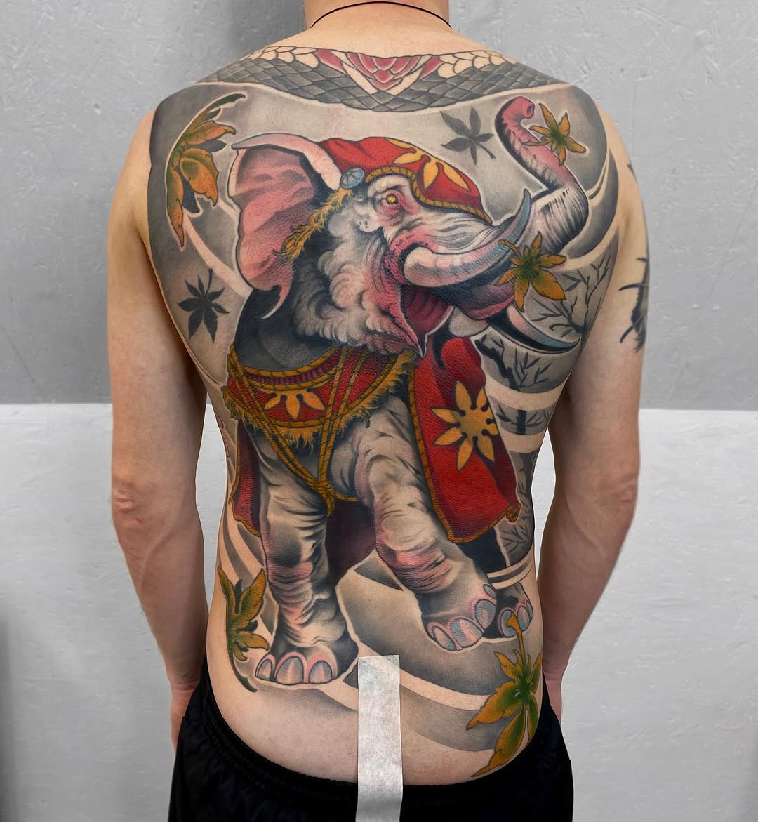 Full-back tattoo of an ornately dressed elephant in red and gold clothing with floral accents, displayed on a shirtless person from behind.