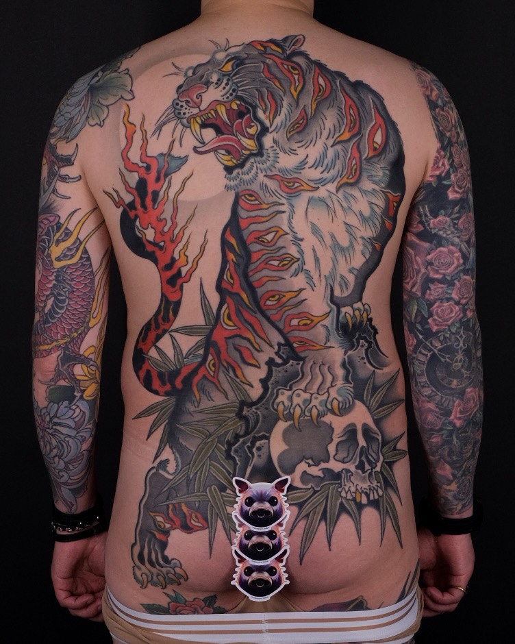 Back tattoo featuring a large tiger head with red and yellow flames, skull, demon face, and floral sleeves on both arms.