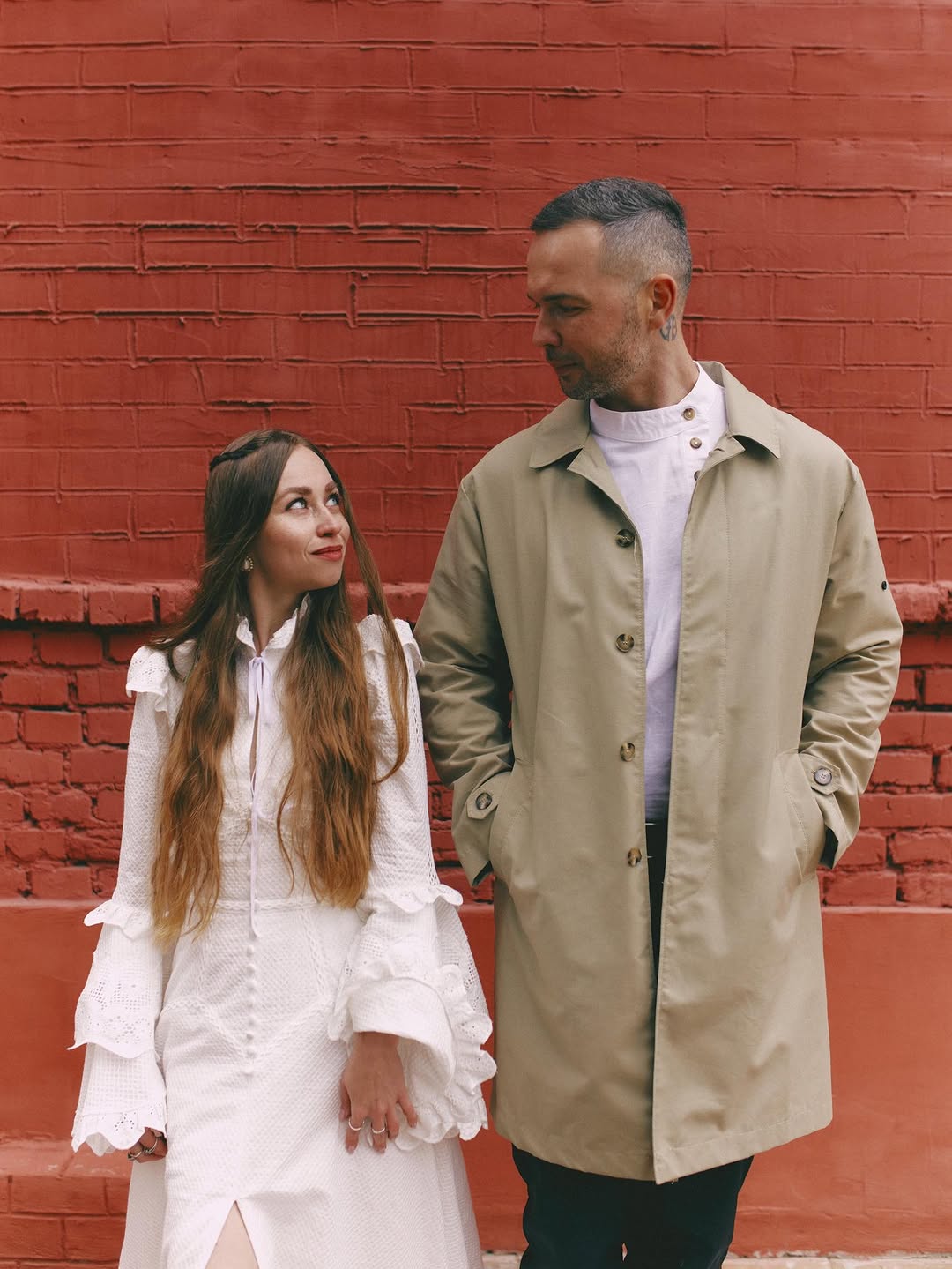 Two people stand against a red brick wall; the man on the right displays a visible tattoo on his neck area in black ink.