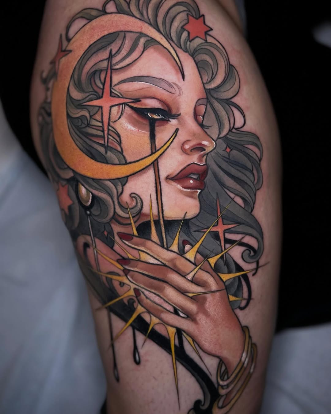 Portrait tattoo of a woman's profile with curled hair, celestial symbols including a crescent moon and crosses, rendered in orange, yellow, and black on the upper leg.