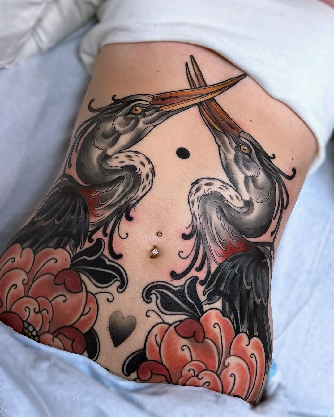 Three herons with long beaks and ornate feathers surround red floral elements on the upper arm and shoulder, rendered in black, gray, and red ink.