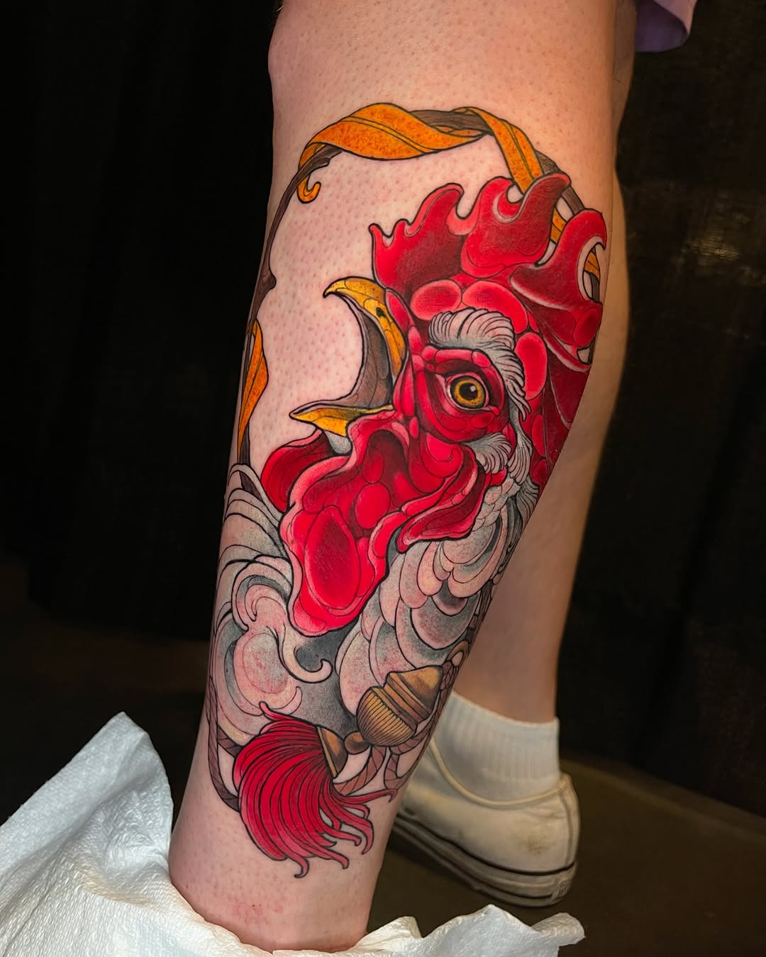 Colorful rooster head tattoo on calf featuring bright red and orange plumage with ornamental scrollwork details below.