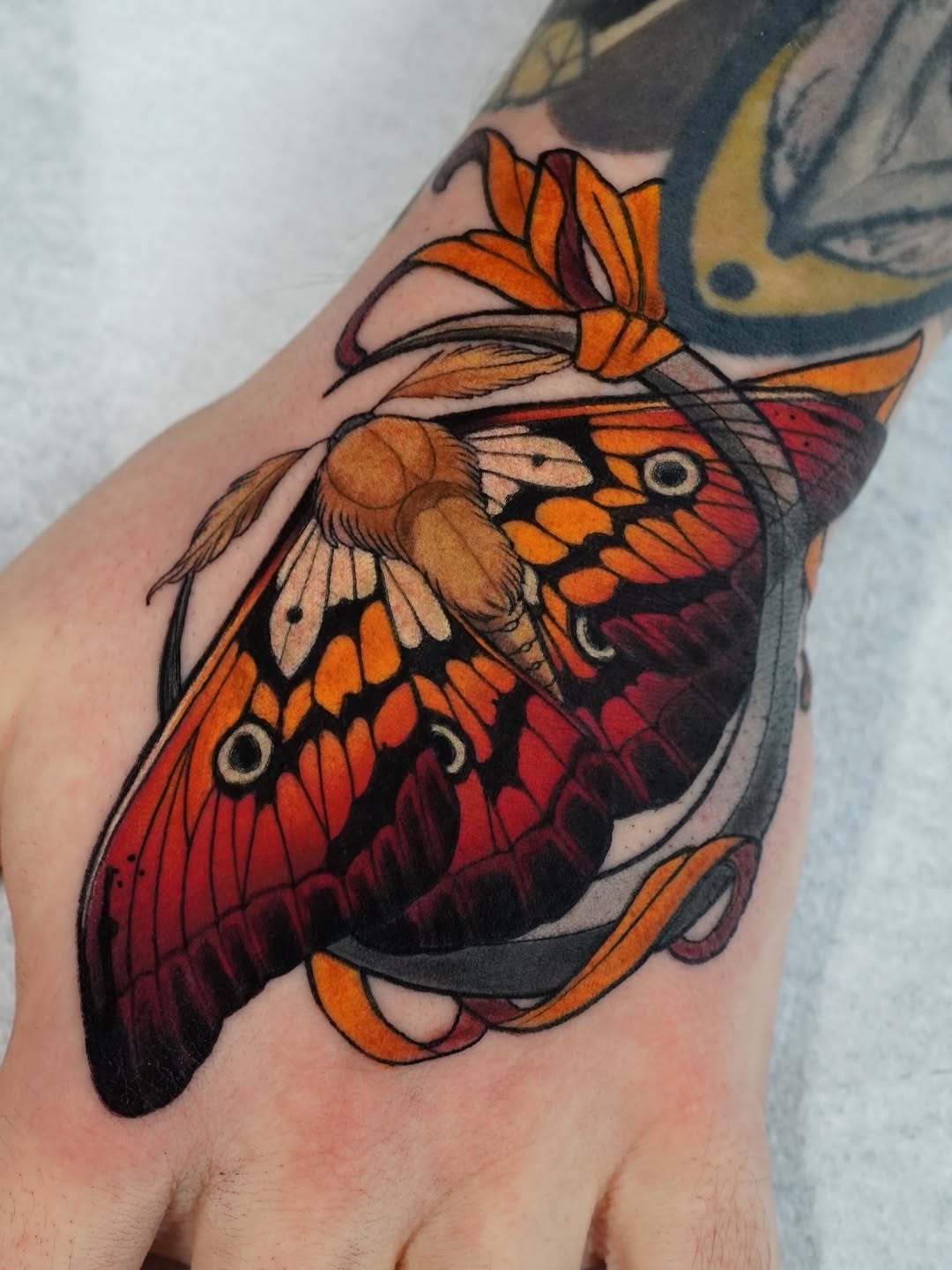 Colorful butterfly tattoo on the hand featuring orange, red, and brown wings with detailed eye spots and black outlines.