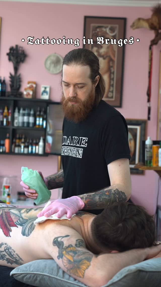 Tattoo artist in pink studio applying a colorful bird design on client's arm using green soap and pink gloves.