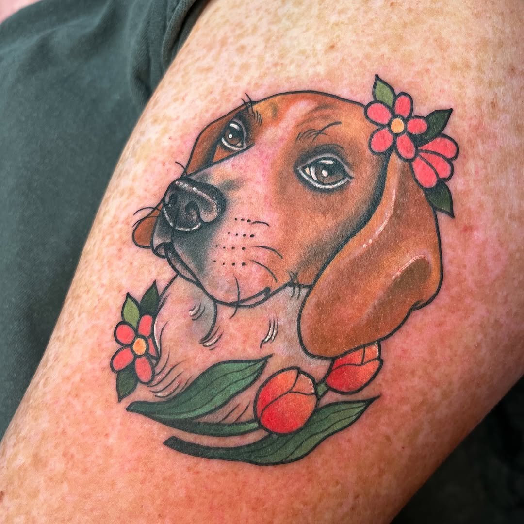 Brown dog's head with expressive eyes adorned with red flowers and green leaves, tattooed on upper arm in red, orange, brown, and green inks.
