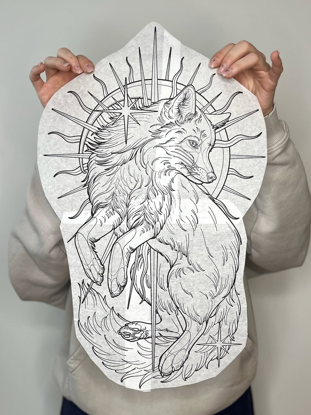 Line drawing tattoo design featuring a wolf head with radiating spikes and a vertical sword through its body, shown on white paper held by hands.