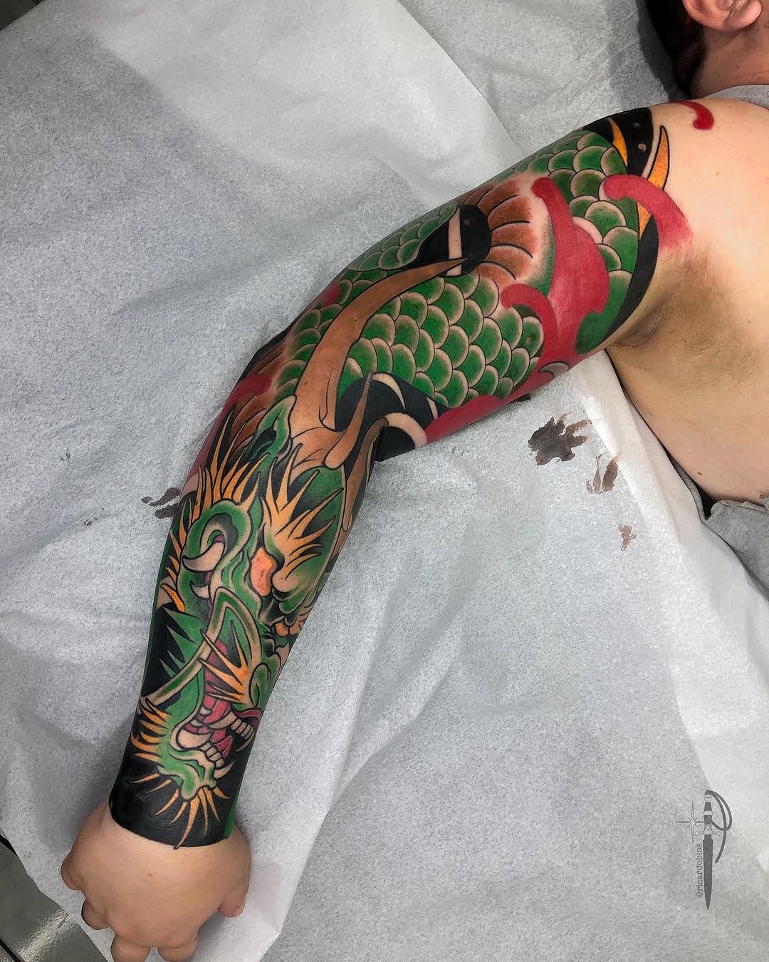 Full-arm sleeve tattoo featuring a large dragon head in green, red, orange, and black with scales, flames, and decorative elements extending from shoulder to wrist.