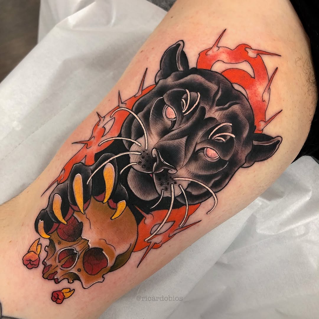 Demon head tattoo with large fangs and horns on upper arm, featuring black shading, red and orange flames, and yellow accents.